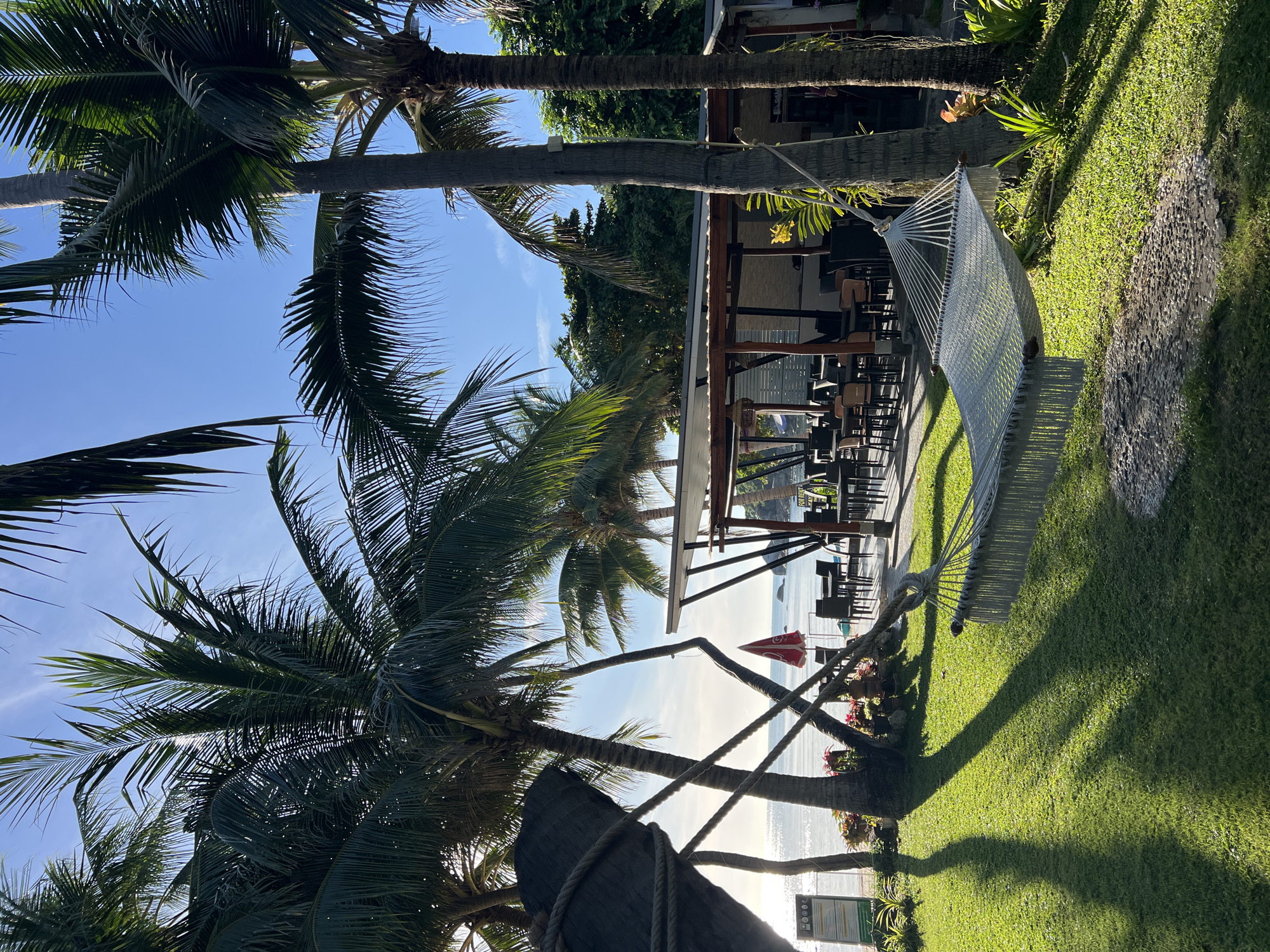 The image depicts a serene tropical scene at Baan Chaweng Noi, Thailand. The foreground is dominated by lush green grass and the shadows of palm trees cast by the bright sun. A white hammock is suspended between two palm trees, inviting relaxation. In the middle ground, an open-air restaurant or bar area is visible, with tables and chairs set up, suggesting a place for dining or socializing. Beyond the restaurant, the calm ocean stretches out under a clear blue sky. The sun's position and the long shadows indicate it is likely late afternoon or early evening. The overall atmosphere is peaceful and idyllic, characteristic of a tropical resort. There is a red umbrella visible near the beach. No visible text is present in the image.