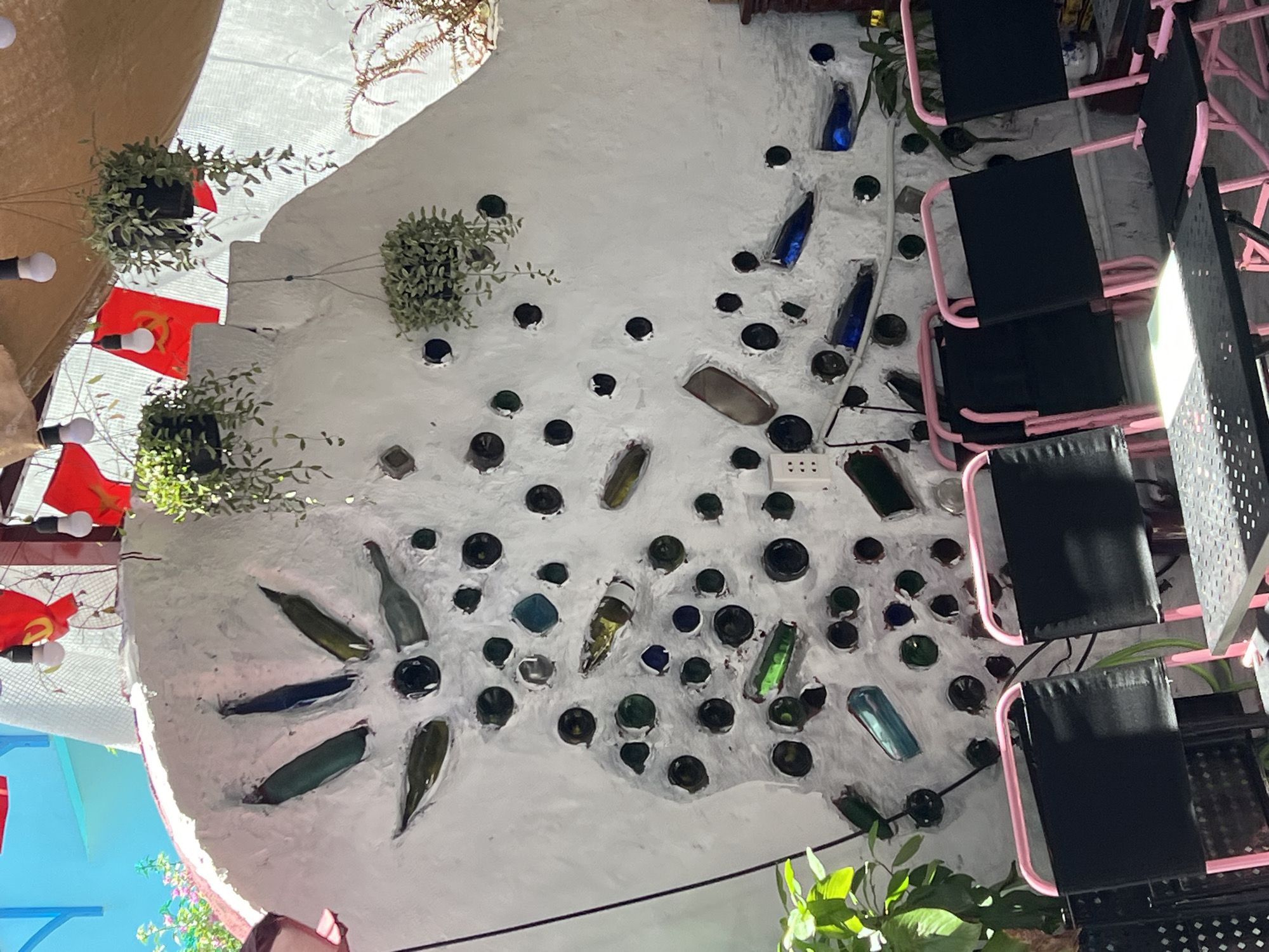 This image depicts a unique wall art installation in the city of Da Nang, Vietnam. The wall, constructed of white concrete, is adorned with embedded glass bottles of various colors, including green, blue, and clear. Many of these bottles are arranged in a sunburst pattern near the top left of the frame, with their necks pointing outward from a central circular element. The wall is also punctuated by numerous circular holes, some of which also contain glass bottles. Several hanging plants with trailing vines are suspended from above, adding a touch of greenery to the scene. In the foreground, a row of black folding chairs with bright pink metal frames are visible, suggesting this is an outdoor seating area, possibly part of a cafe or restaurant. Red flags, possibly political or decorative, are also visible hanging in the background, as are several pendant light bulbs. The overall impression is a creative and somewhat rustic decorative feature integrated into an outdoor urban setting.