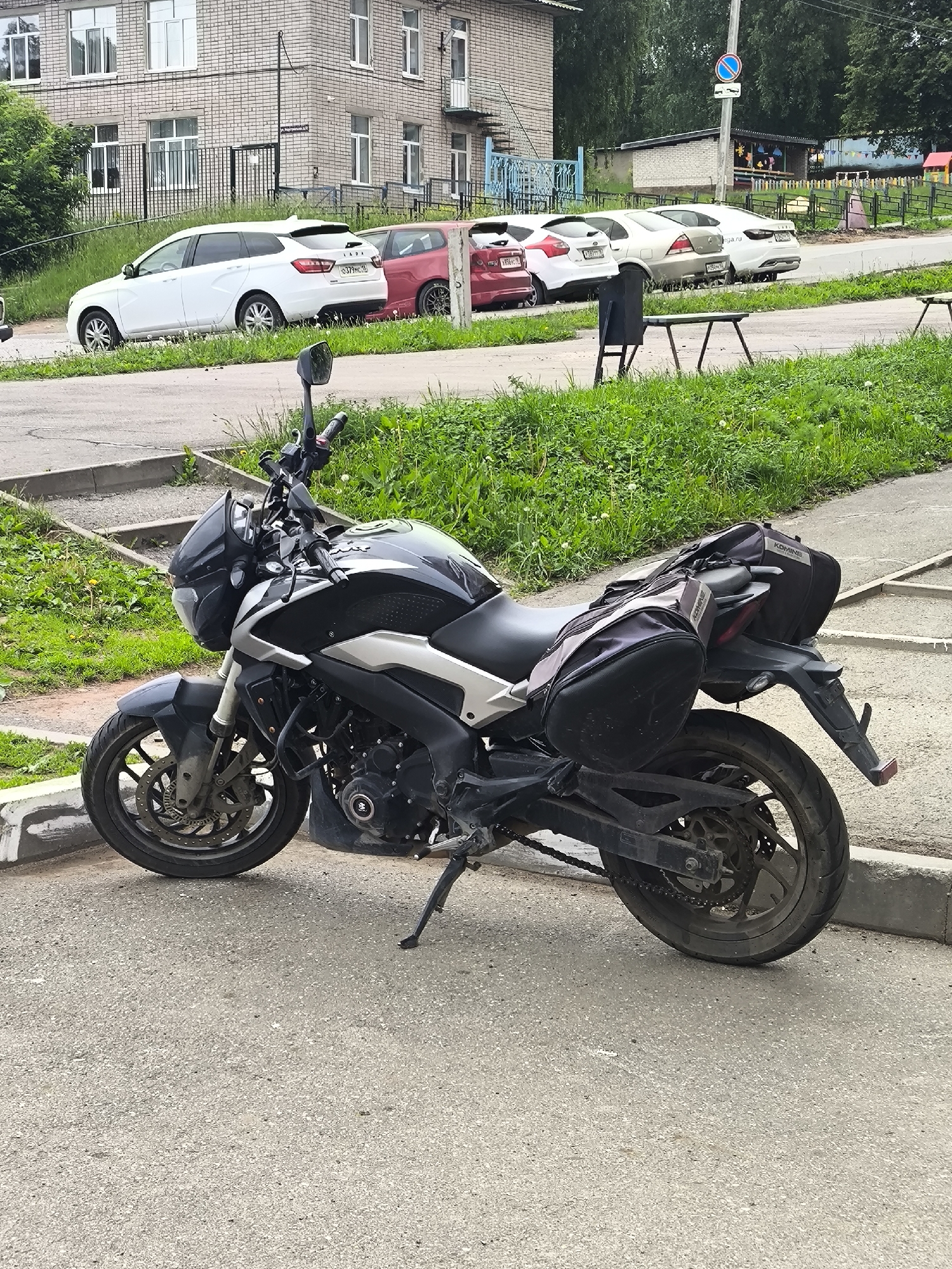 This image depicts an outdoor urban scene in Izhevsk, Udmurt Republic, Russia, identified by the "18" region code on visible vehicle license plates.
In the foreground, a black and silver Bajaj Dominar motorcycle is parked on an asphalt road, facing left, supported by its kickstand. The motorcycle is equipped with "KOMINE" brand soft saddlebags mounted on the rear and a tank bag on the passenger seat. The front brake disc and caliper show some accumulation of dirt. The asphalt road surface is light gray and speckled.
Behind the motorcycle, a concrete curb separates the road from a grassy verge featuring green grass and scattered dandelions.
In the midground, several parked cars are visible along the road. From left to right: a white Lada Vesta SW Cross (license plate O379ME 18), a partially visible red car, a silver sedan (partially visible license plate P50_T 18), and a white sedan (partially visible license plate E_0T_ 18). Further left and partially cropped, another red hatchback (license plate T556__ 18) is visible.
In the background, on the left, stands a multi-story brick building with a light brown/beige facade, featuring numerous white-framed windows and balconies. A black metal fence runs in front of this building. To the right, a blue circular road sign depicting a white truck with a downward arrow is mounted on a tall dark pole. Further right, a small black metal trash receptacle and a weathered wooden table with metal legs are visible. Beyond the parked vehicles, a small, single-story brick structure with a dark roof is present, followed by a colorful children's playground area enclosed by a fence. Dense green trees and foliage form the furthest background. The lighting suggests an overcast sky, with no strong shadows. No individuals are present in the frame.