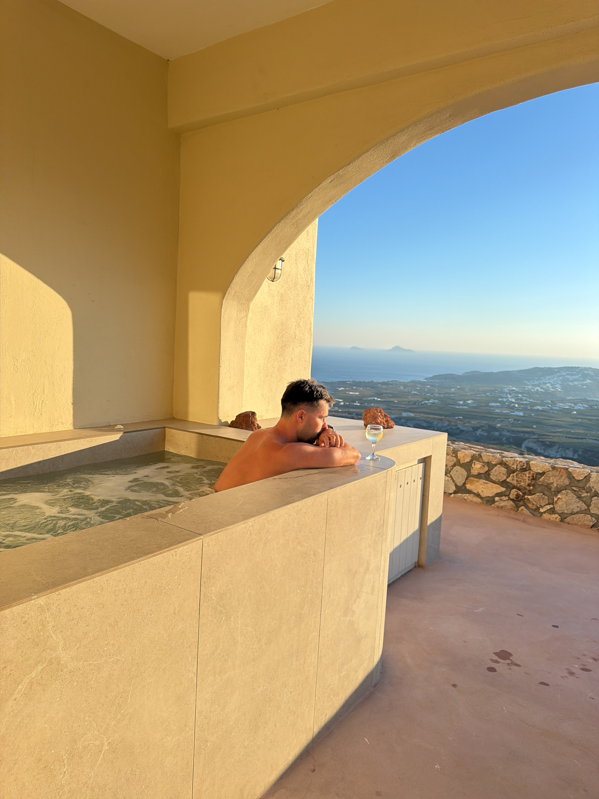 A man with dark hair is partially submerged in a hot tub, leaning on the edge and looking out at a vast seascape with islands and white-washed buildings. He is wearing a watch on his left wrist. A glass of white wine sits on the edge of the hot tub beside him. The hot tub is constructed from light-colored, tiled material. The scene is set on a terrace with a stone wall bordering a pinkish-brown floor. Above, a pale yellow archway frames the sky and the distant view, suggesting an outdoor setting on a clear day. The sunlight casts long shadows across the terrace. The location is likely an upscale villa or hotel in Thira, Greece, given the architectural style and the characteristic landscape of the Cycladic islands.