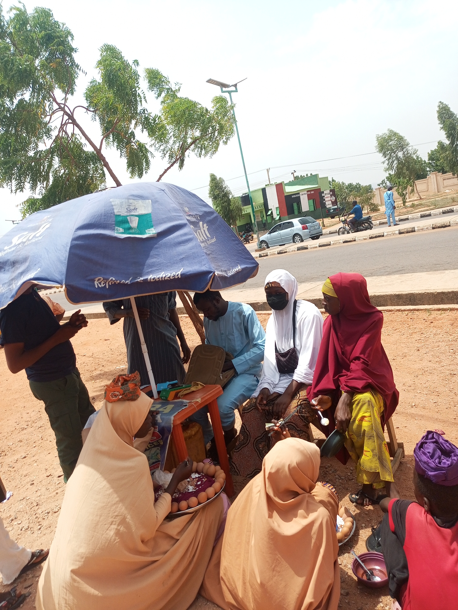 A group of people is gathered around a small orange table shaded by a blue umbrella. One person in a peach-colored hijab holds a plate of eggs. Several others are seated, and some are standing. The setting appears to be outdoors, near a street with vehicles passing by and buildings in the background. A light pole stands near a tree.