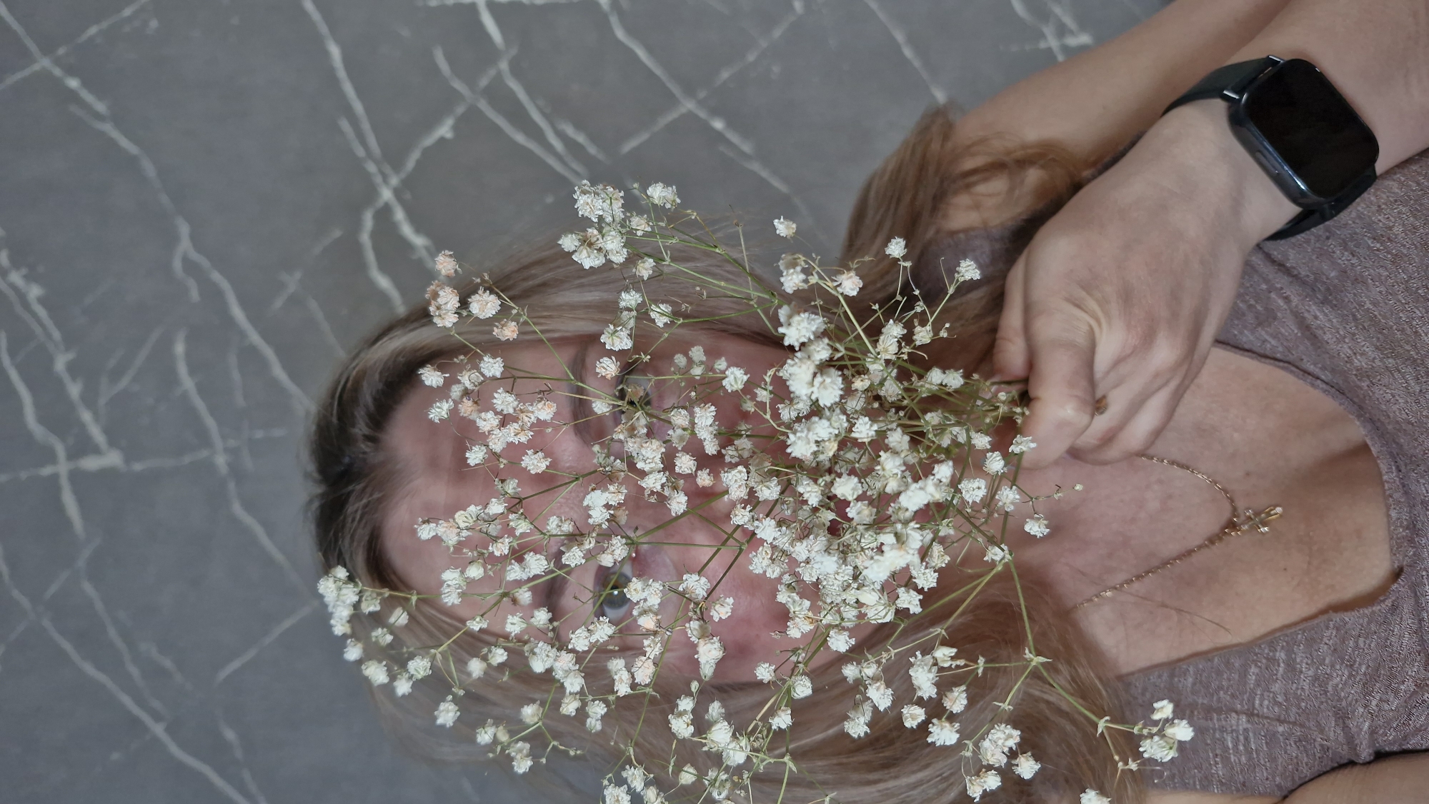 A middle-aged woman lies against a gray marbled surface. She is holding a sprig of baby's breath flowers in front of her face, obscuring most of her features. Her eyes are visible through the flowers. She is wearing a brown top and a gold necklace with a cross pendant. A smart watch is visible on her left wrist. The location is unspecified; there is no indication of the city of Otchor, Russia.