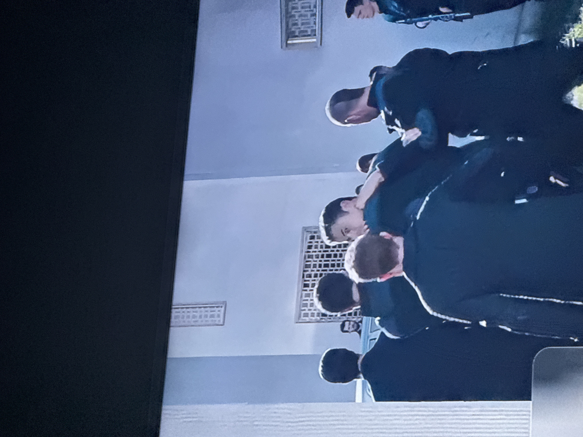 A group of individuals dressed in dark uniforms are standing indoors. The location appears to be a building in the city of Mytishchi, Russia, as suggested by the architectural style and the presence of ventilation grids in the wall. The people are positioned in a manner that suggests they are either bowing, or bending over. The setting appears to be institutional, possibly a school or governmental building.