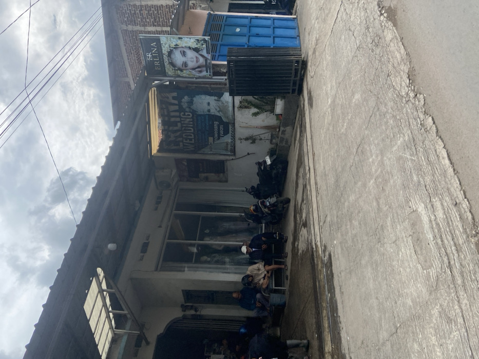 The scene is in front of a storefront in Ciparay, Indonesia, located at coordinates -7.023 latitude and 107.688 longitude. The storefront is a small, white building with a large window displaying mannequins wearing dresses. A sign above the window advertises "ERLINA WEDDING" in large letters. Next to the storefront is a blue metal gate. There are three men sitting on the sidewalk in front of the storefront. Two of the men are wearing white hats. Two motorcycles are parked in front of the storefront. The sidewalk is paved with concrete and there are cracks in the pavement. The sky is overcast and gray. The environment is urban.