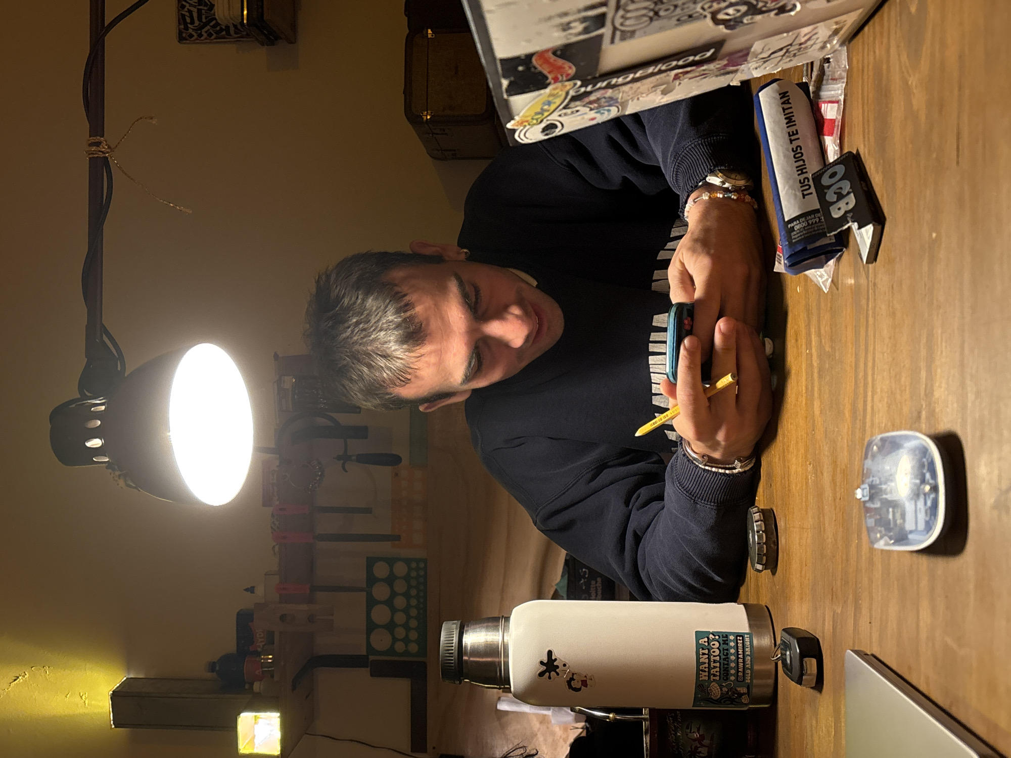 A person is seated at a wooden table under a bright desk lamp. They are wearing a dark blue long-sleeved shirt and have several bracelets on their wrists. The person is focused on an object they are holding in their hands, using a yellow pencil. On the table, there's a water bottle with stickers, a car key, a small electronic device, a laptop, and a box with "TUS HIJOS TE IMITAN" printed on it. Various crafting tools are hanging on the wall behind the person. The brand "OCB" is visible on an item on the table. While the location of the city of Buenos Aires, Argentina is not directly apparent in the image, there is a reference on the box with "TUS HIJOS TE IMITAN" which includes an Argentina help line.