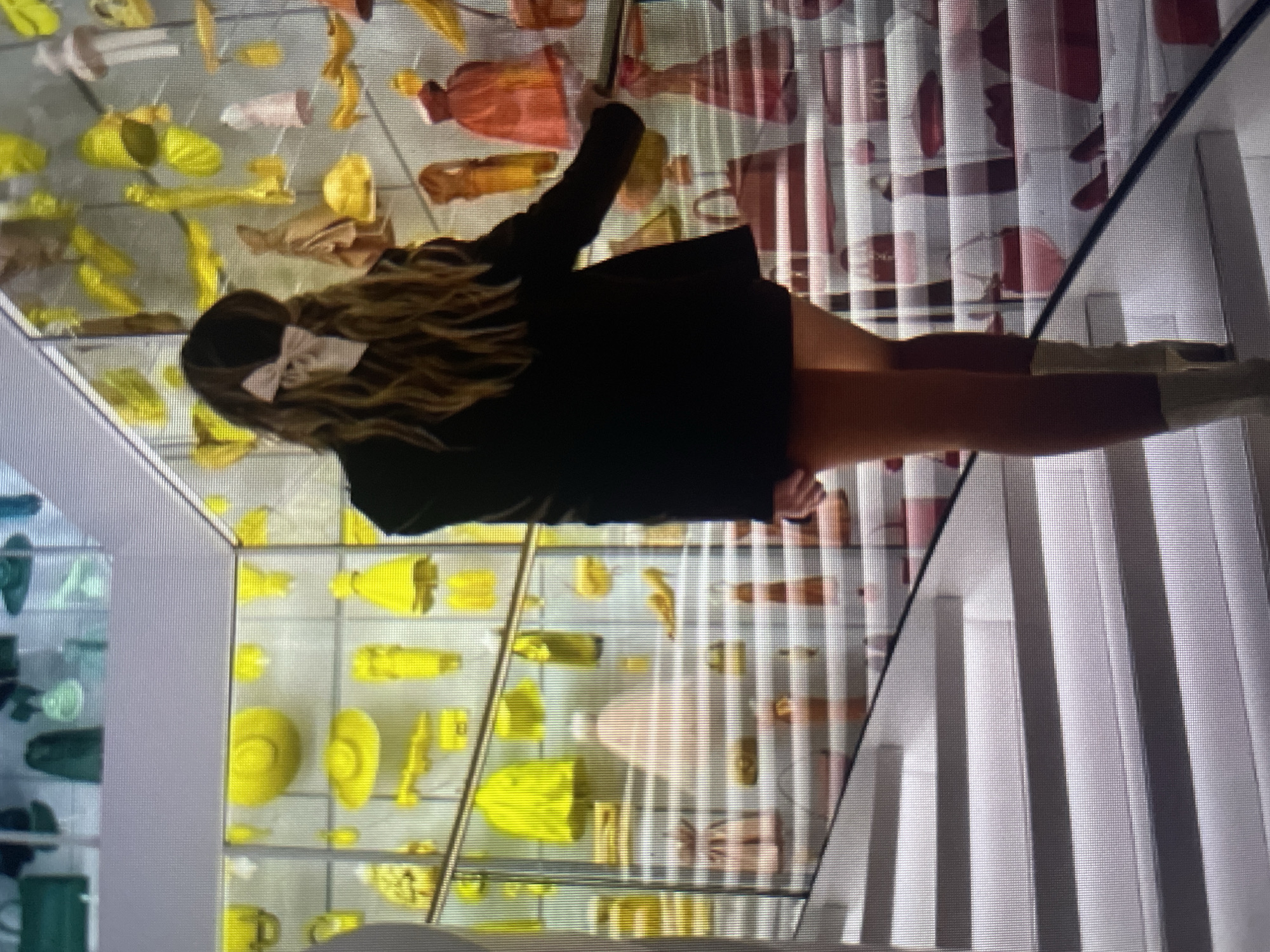 In an indoor setting, likely a museum or exhibition, a person is ascending a modern, white staircase. The person, viewed from behind, has long, wavy brown hair adorned with a light pink bow. They are wearing a dark blazer over a short skirt and light-colored boots. The background features a display wall filled with a vibrant collection of yellow and pink fashion items, including hats, purses, and dresses, artfully arranged in illuminated glass cases. The stairs themselves are also adorned with fashion accessories, primarily pink and red purses and shoes, creating a visually striking presentation. The scene is brightly lit, emphasizing the colorful displays and the polished surfaces of the architecture. The city of Kołobrzeg, Poland, is not depicted in this image.