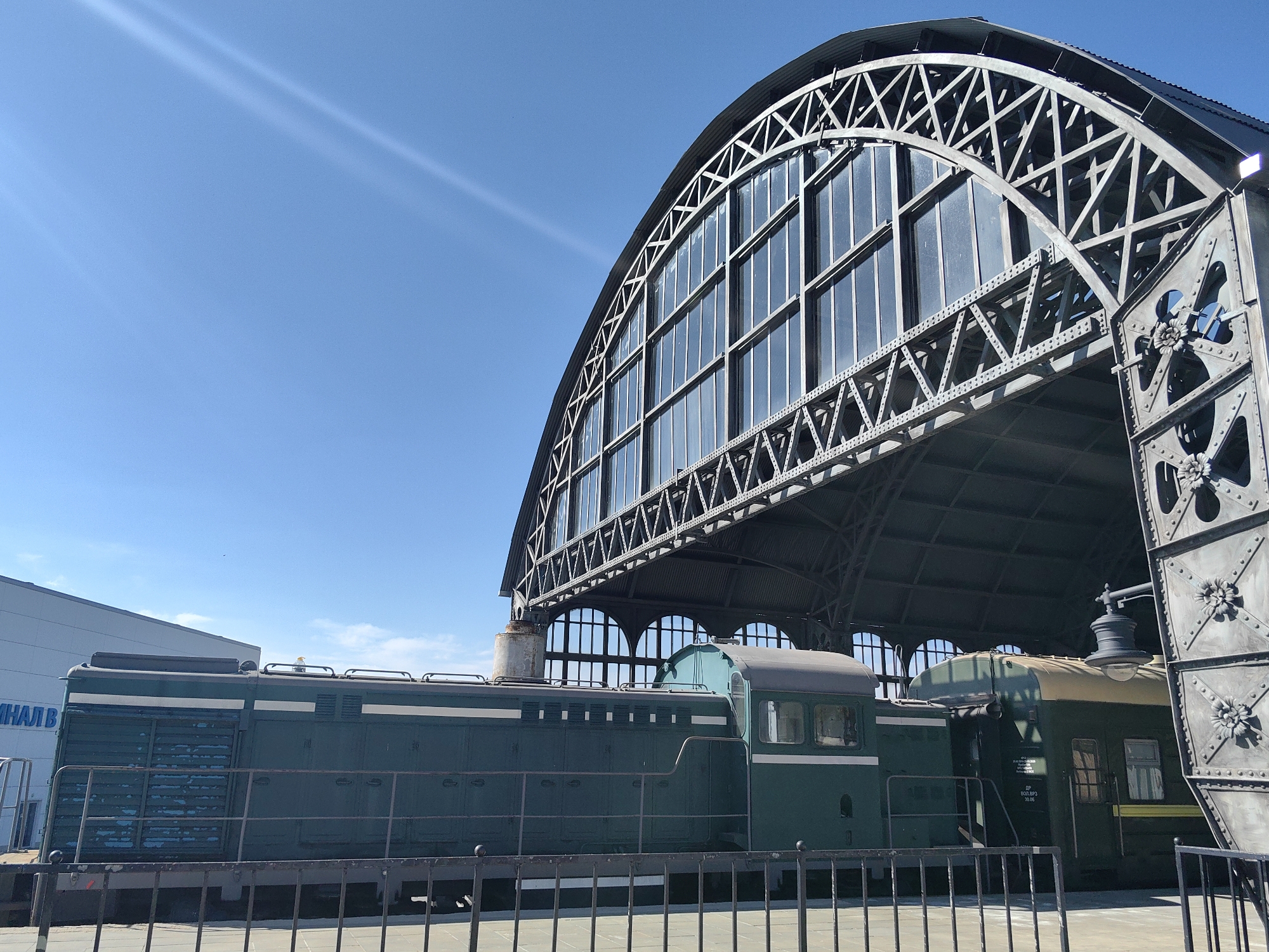 A low-angle outdoor shot captures a green train engine parked beneath a large, arched train shed with a steel truss structure and glass panels. The train shed's roof is a dark, corrugated metal. In the background, a modern building with the partial Cyrillic text "МНАЛ В" is visible on its side. The sky is clear and bright blue with a faint contrail. The train engine itself has a dark green exterior with lighter green trim and a series of vents along its side. A black metal fence runs in front of the train.