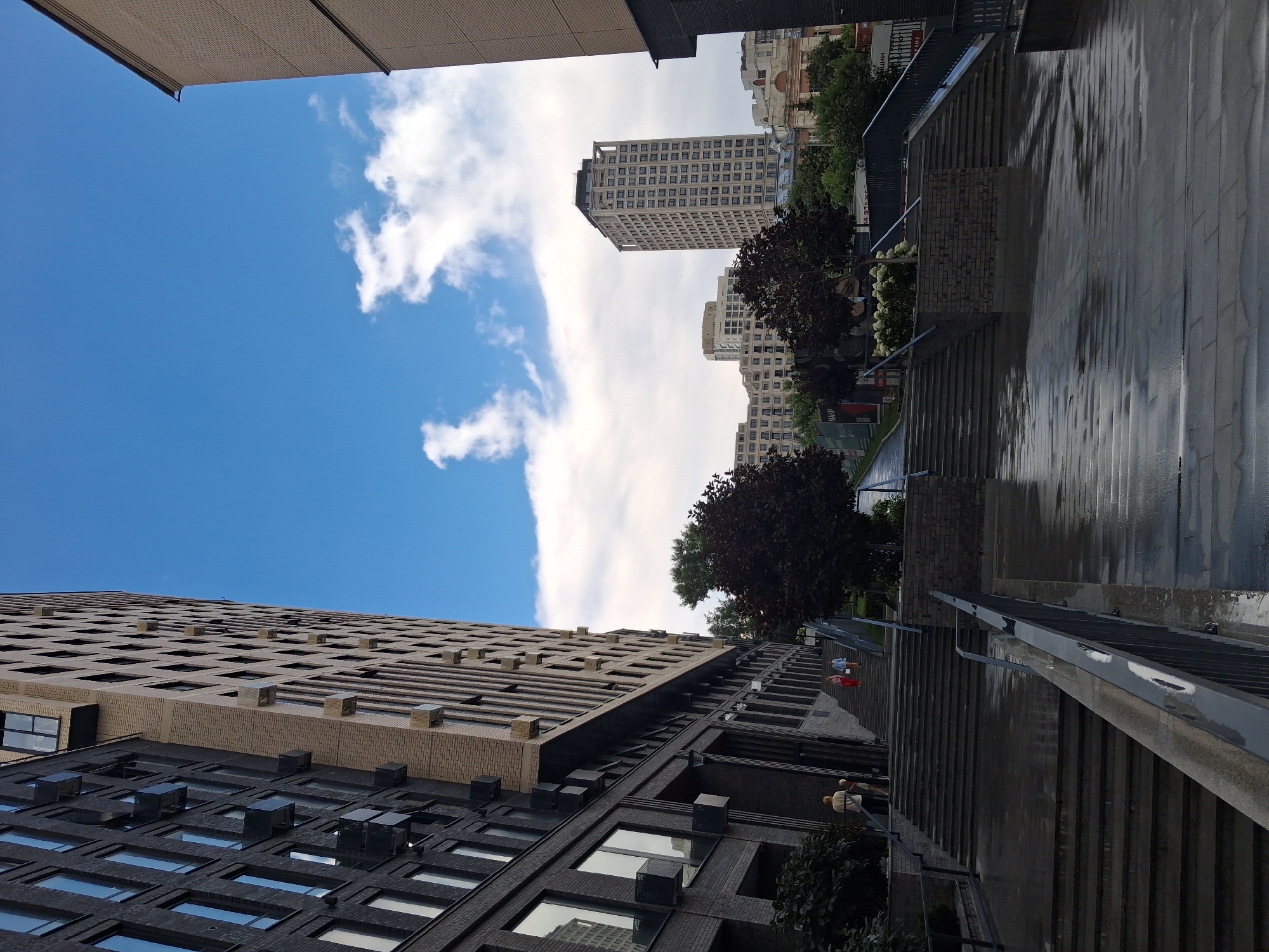 The image depicts an outdoor scene in front of modern apartment buildings. A wide, wet concrete staircase leads upwards, flanked by dark metal railings. To the left, a tall building with a dark facade and numerous windows dominates the foreground, angled into the frame. To the right, another building with a lighter facade is partially visible. In the mid-ground, more apartment buildings of varying heights and architectural styles are visible, with trees and greenery interspersed. The sky above is blue with scattered white clouds. A few people can be seen ascending the staircase. The scene appears to be a public outdoor space, possibly a courtyard or plaza, between residential buildings.
The location is the residential complex "Sozvezdie" (Constellation) in Novosibirsk, Russia.