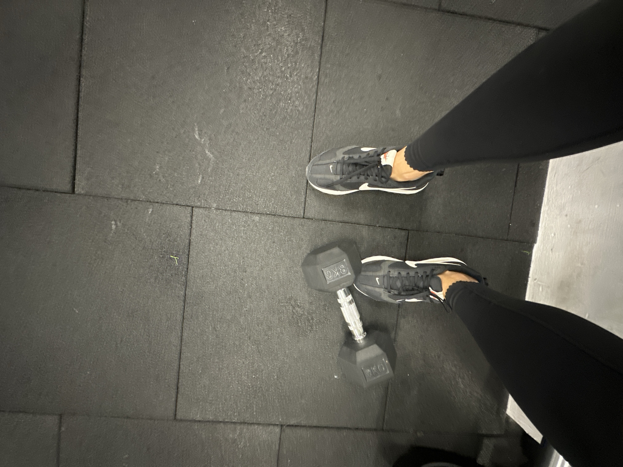 The image shows a close-up view of a person's legs and feet on a dark gray, tiled gym floor. The person is wearing black athletic leggings and black Nike sneakers with white accents. One foot is positioned close to an 8kg dumbbell, which is also dark gray and hexagonal in shape, with "8KG" clearly visible on its sides. The other foot is further away, and its sneaker is also visible. The scene suggests the person is either about to start or has just finished a weightlifting exercise. The lighting is dim, typical of an indoor gym environment. The tiled floor and the presence of weights are strong indicators of a gym setting. There are no explicit indicators of time of day, weather, or emotions. The only visible text is "8KG" on the dumbbell.