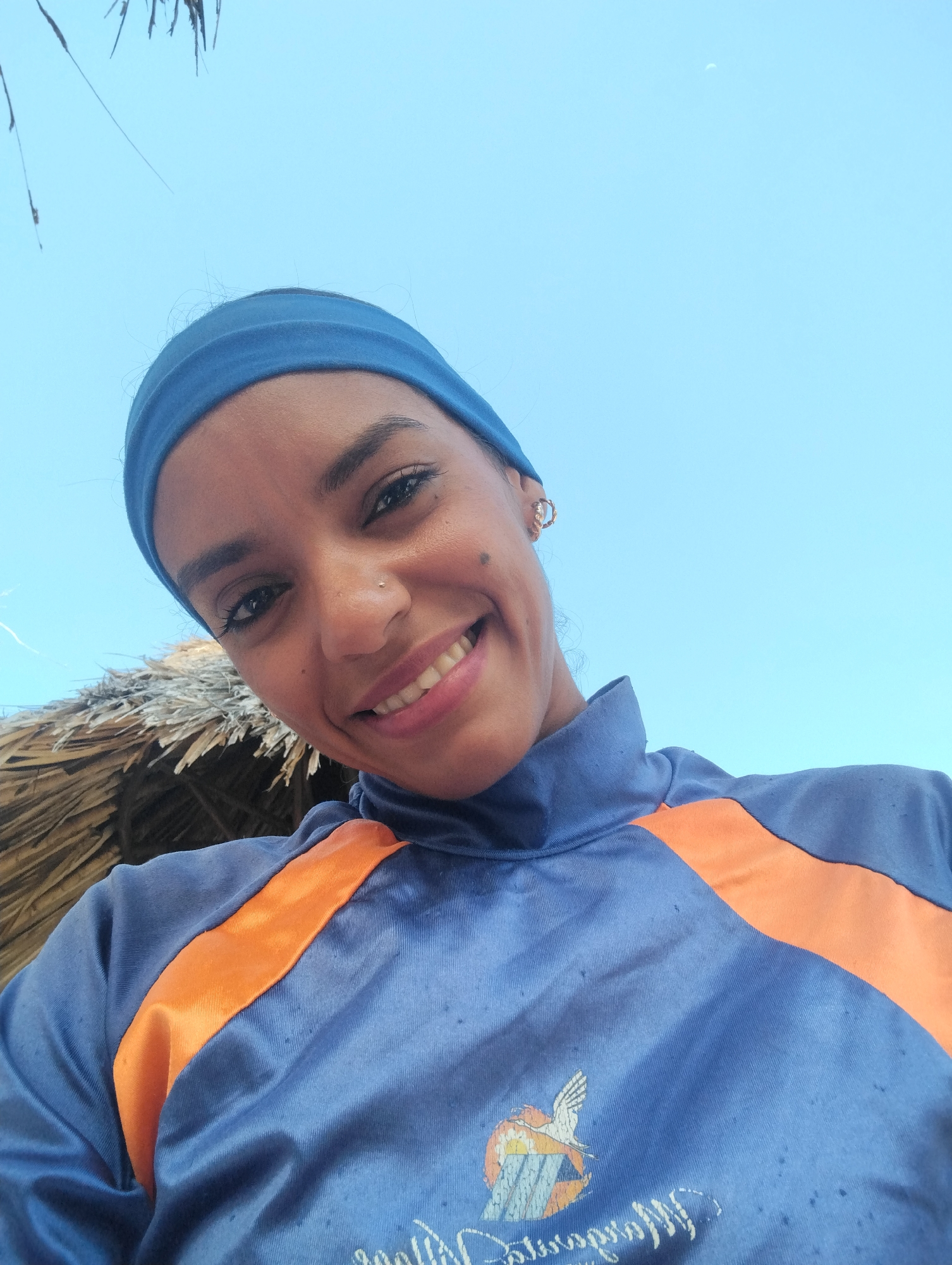 A smiling woman with a blue headband and a blue and orange shirt poses for a selfie. She has dark hair, eyebrows, and eyelashes. A nose ring is visible. Behind her is a blue sky and the edge of a straw roof. The emblem of the Universidad Corporativa Sigo can be seen on her shirt. This image might have been taken in Porlamar, Venezuela, given that Universidad Corporativa Sigo has locations there.