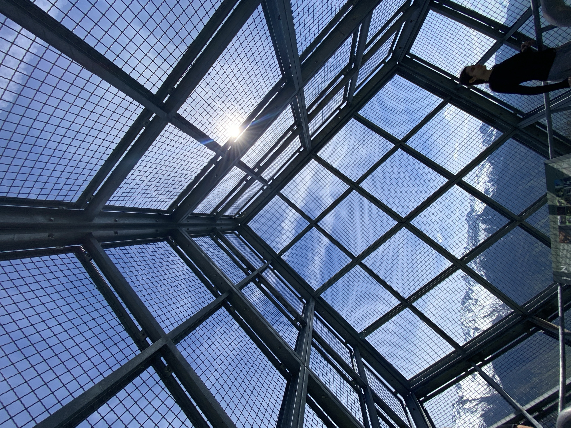 The image captures a striking upward view through a modern, industrial-style observation structure, likely a skywalk or viewing platform in Saint-Georges-d'Hurtières, France. The primary subject is a dark, sturdy metal framework forming a dense grid pattern across the entire ceiling and upper walls. Through the square openings of this grid, a bright blue sky with scattered white clouds is visible.
The sun is prominently featured in the upper-left quadrant, shining brightly through the grid and creating a distinct sunburst and lens flare effect. In the lower-right section, a person is partially visible, appearing to stand or lean against a barrier, looking upwards or outwards. They are dressed in dark clothing, silhouetted against the bright background.
Reflected in or seen through the transparent panels beneath the metal mesh, a majestic snow-capped mountain range or large rocky outcrop is discernible, suggesting a high-altitude or alpine setting. The strong sunlight indicates it is daytime, and the clear skies point to fair weather. The low-angle perspective emphasizes the imposing structure and the vastness of the sky above. On the far right, a partial view of a sign or display board shows fragments of text, including "MMAGATE" and "DMORE". The combination of the rigid architectural lines and the natural grandeur of the mountains creates a dynamic and immersive scene.