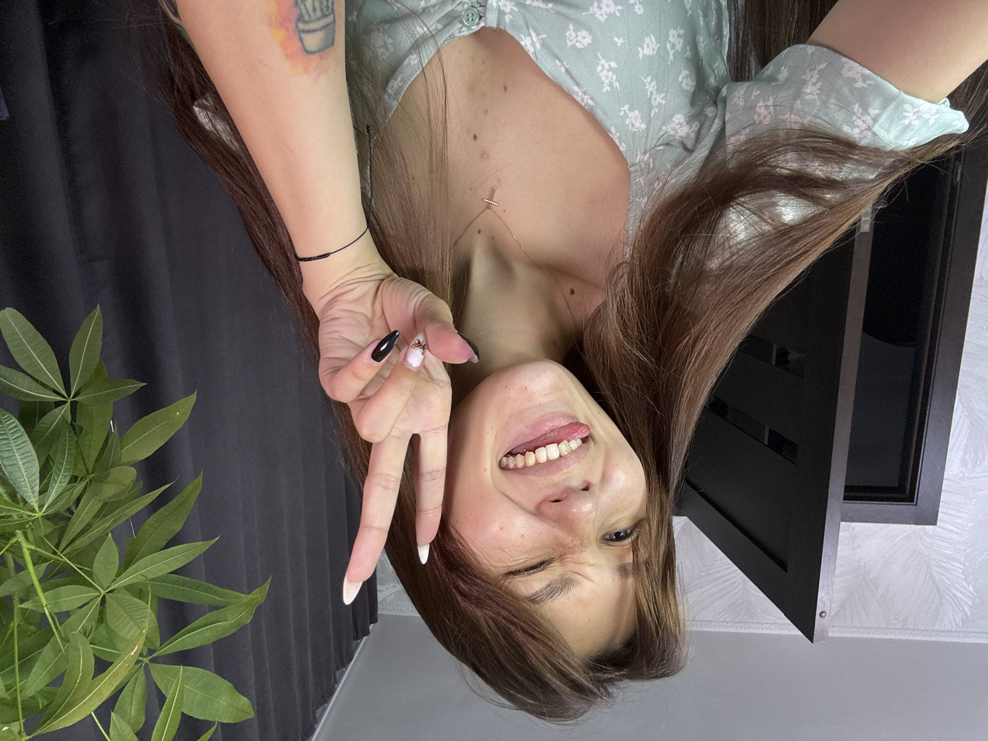A young woman is taking a selfie in a room. She has long brown hair and is wearing a light green floral dress. She is winking and sticking her tongue out, while holding up two fingers in a peace sign. Her right arm is bent at the elbow, and her hand is near her face. She has a cross necklace on. Her left arm is extended, and she has tattoos on her forearm. A large green plant is visible to her right, and a dark curtain is in the background. A door and wallpaper are visible to her left. The scene appears to be indoors, likely in a residential setting. There is no specific information to indicate the location is in Novokuznetsk, Russia.