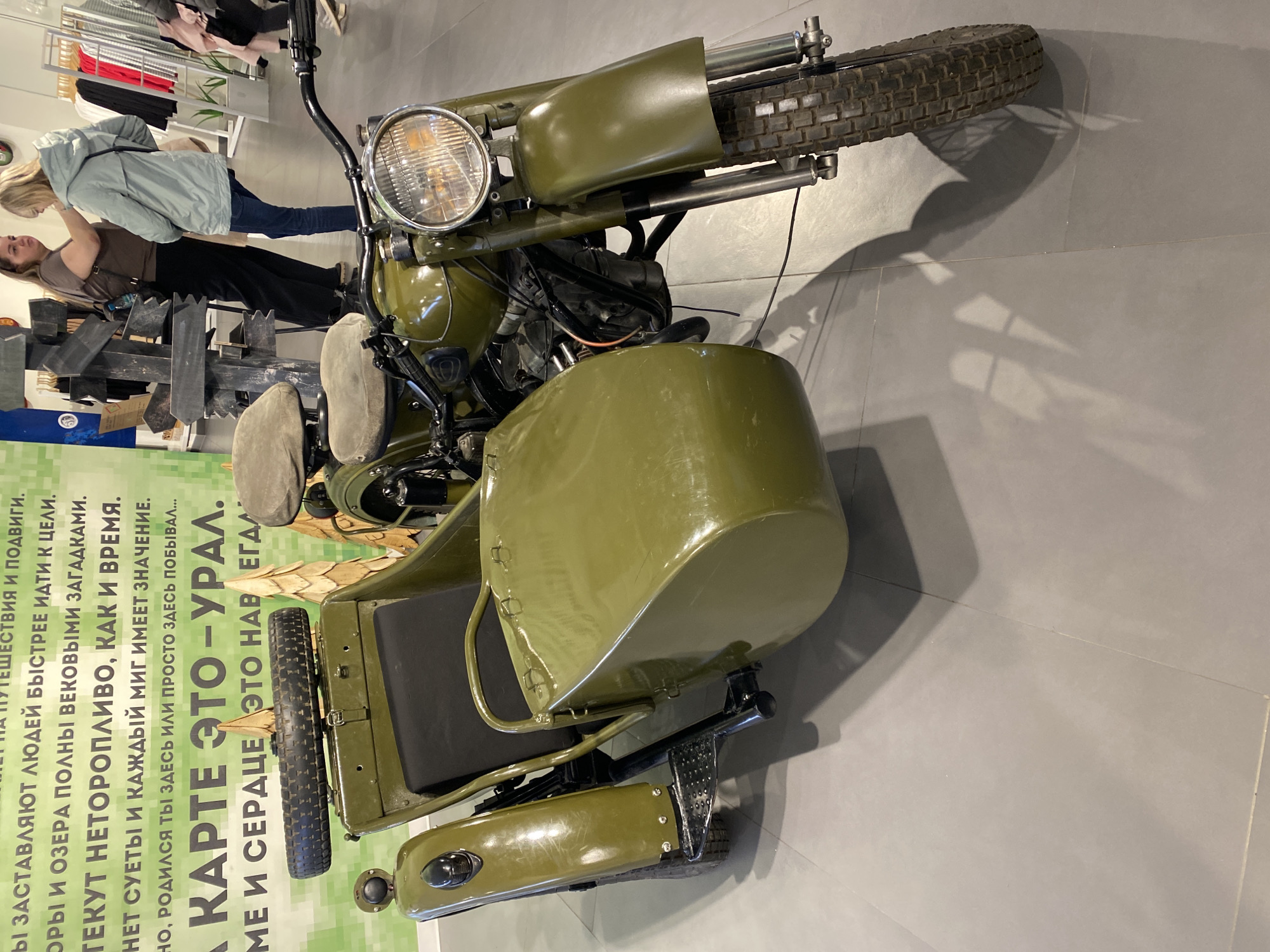 The image depicts an olive green motorcycle with a sidecar. The motorcycle has a large headlight and knobby tires. The sidecar is attached to the right side of the motorcycle and has a wheel, a seat, and a fender. The motorcycle is located indoors, possibly in a shopping mall or a showroom. In the background, people can be seen browsing through clothes on display racks. To the left of the motorcycle, a green sign with text in Russian is partially visible. Based on the text "КАРТЕ ЭТО - УРАЛ," and "родился ты здесь," the motorcycle is likely an Ural model and the location is in or related to the Ural region, which includes Yekaterinburg, Russia.