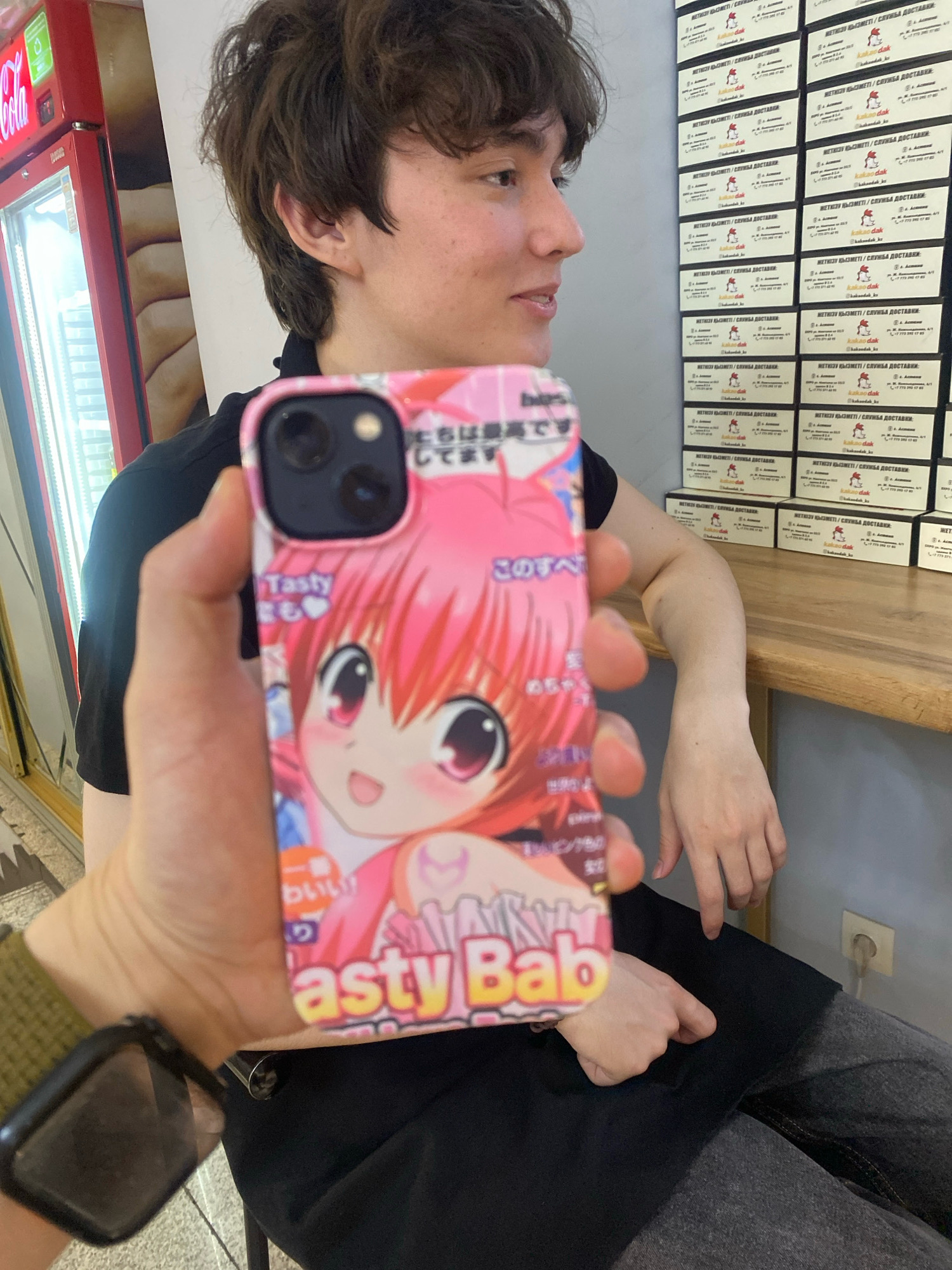 A young person with brown hair and light skin is in the foreground of the image. They are wearing a black t-shirt and grey pants. The person is holding up a smartphone with a pink case featuring an anime character. The person's right arm is resting on a wooden table. In the background, there is a red vending machine and a wall covered with business cards. The scene appears to be indoors, possibly in a cafe or shop. The image is taken from a slightly low angle.