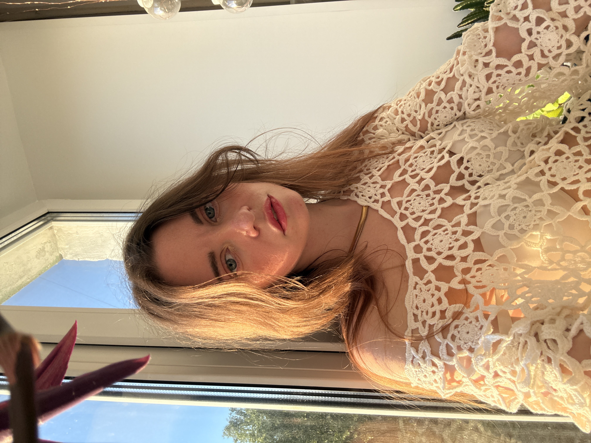 The original image, rotated 90 degrees counter-clockwise, depicts a young woman in an indoor setting, viewed from a low angle looking upwards. She has long, light brown hair, which is illuminated by warm, natural light, and light-colored eyes (appearing blue or green). Her lips have a soft pink hue. She is wearing a cream or off-white crocheted garment, possibly a top or shawl, featuring intricate floral patterns, and a thin golden necklace is visible around her neck.
The woman is positioned near a white window frame, and through the window, a clear blue sky and the upper parts of green trees are visible, indicating a sunny daytime scene. The sunlight streams in, creating highlights on her hair and the crocheted fabric. Above her, a plain white ceiling with small, globe-like light fixtures can be seen. In the blurred foreground, towards the bottom left, are the dark red or purple leaves of a houseplant.
The woman is posing for the camera, looking directly at the viewer with a calm and serene expression. The overall atmosphere is warm and gentle, largely due to the soft, natural lighting, suggesting it might be either early morning or late afternoon (golden hour). There is no visible text in the image. The location context provided for the image is Chaikovsky, Russia.