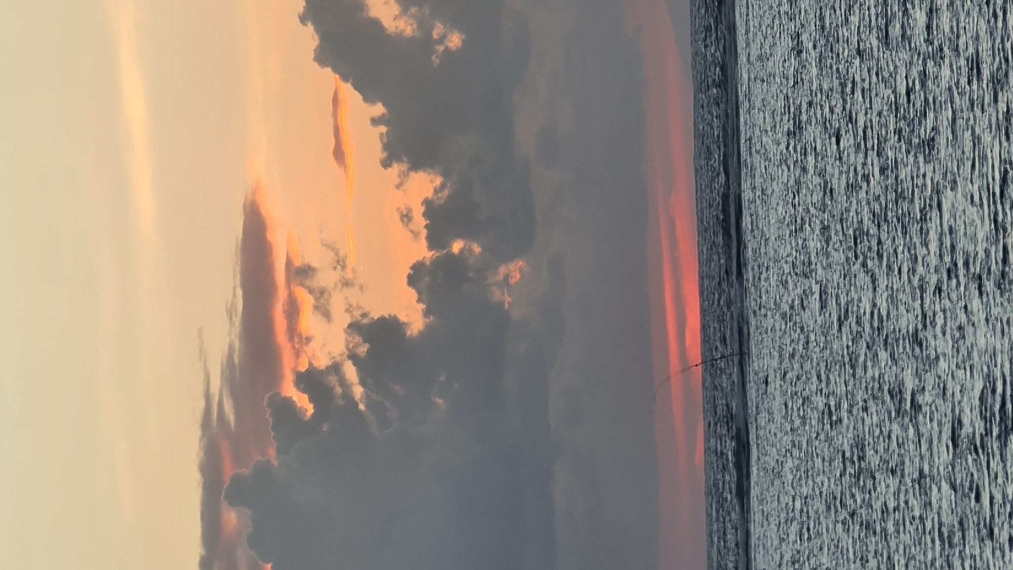 An outdoor, vertical, medium shot captures a sunset over the ocean in Denpasar, Indonesia. The sky is a gradient of soft orange and yellow, with dark grey clouds tinged with pink. The ocean, in the foreground, is dark and choppy with visible waves. A single, thin fishing pole extends from the water into the sky, angled to the left. The horizon is a thin line of red light from the setting sun.