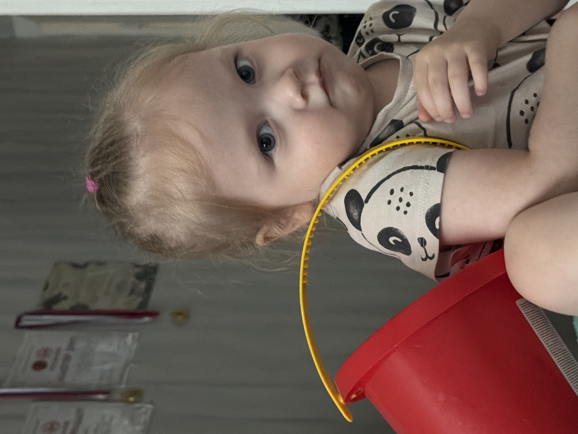 A young child, appearing to be a toddler with light blonde hair pulled back in a small bun with a pink elastic, is centrally positioned and looks directly at the viewer with light blue eyes and a neutral to slightly serious expression. The child is wearing a light tan or beige short-sleeved t-shirt patterned with black stylized panda or bear faces. A bright red plastic bucket is visible in the lower-left foreground, with a textured yellow plastic strap, likely the bucket handle, draped over the child's right shoulder (viewer's left). The child's left hand rests on their chest, partially obscured by the yellow strap. The background consists of a plain light grey wall, upon which several out-of-focus framed items or certificates are hanging, some appearing to have red ribbons and gold medals. No specific geographic identifiers linking the scene to the city of Perm, Russia, are discernible within the image.