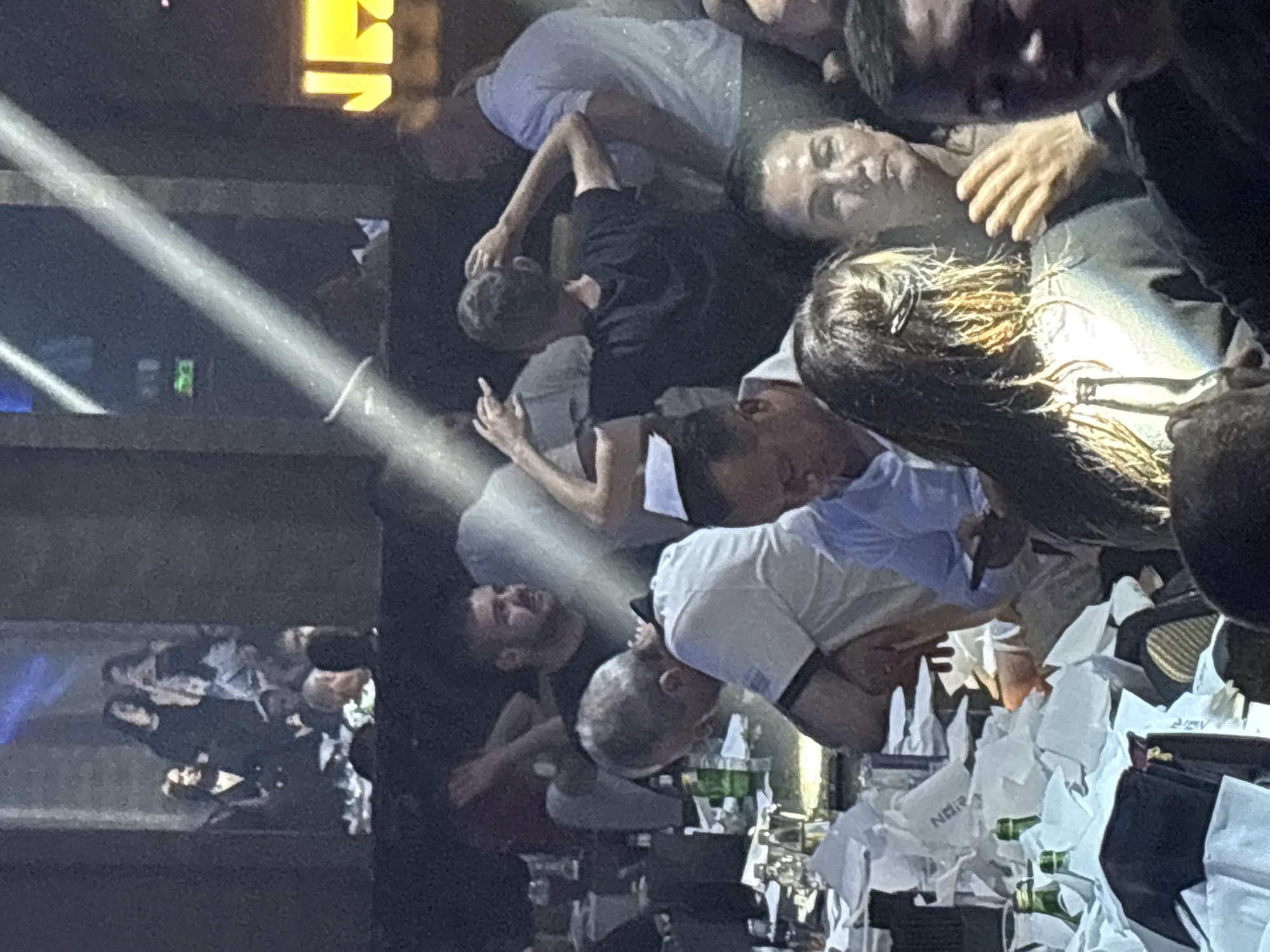 The image captures a crowded scene, potentially inside a nightclub or a similar venue in Unknown, Unknown. The lighting is dim, with a spotlight shining through the center of the frame. A group of people are clustered together, some interacting with each other, while others appear to be looking in different directions. There are several people dressed in casual attire such as t-shirts and collared shirts. A table is visible with numerous empty bottles and crumpled napkins on it. The overall atmosphere suggests a social gathering in a late-night setting.