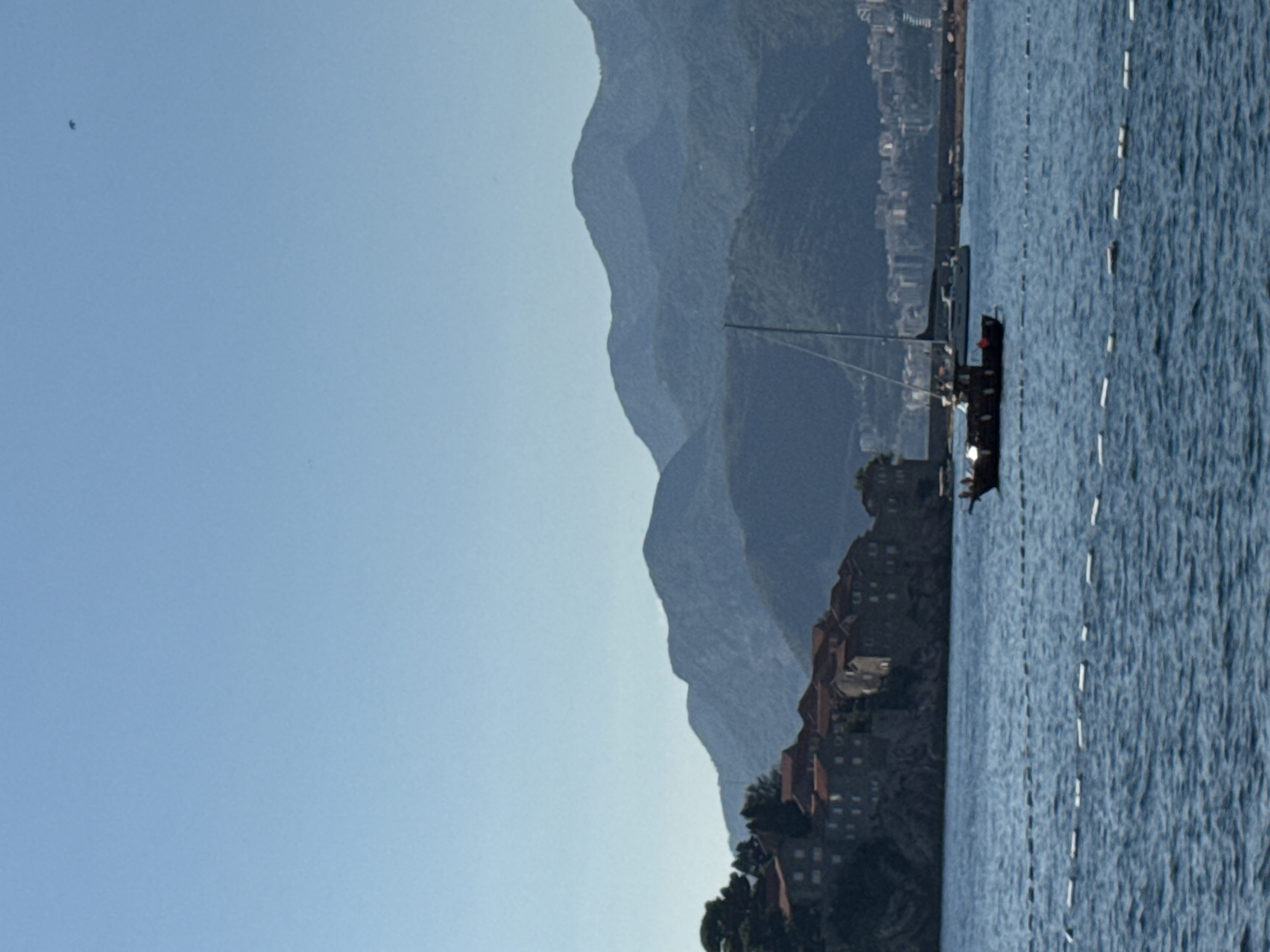 This image depicts a scenic view of Sveti Stefan, Montenegro, featuring a cluster of buildings with red-tiled roofs situated on a rocky outcrop. The buildings appear to be an old village or town, with several structures visible, showcasing a traditional architectural style. The outcrop is surrounded by calm, blue water, suggesting it might be an island or a peninsula connected by a narrow causeway, which is not fully visible.
In the foreground, the water is a deep blue and shows some ripples, indicating a gentle breeze. A line of white buoys or markers stretches across the water, possibly delineating a swimming area or a navigation channel.
Several boats are present in the water. A dark-colored boat, possibly a motorboat or a small yacht, is closer to the foreground, with several people visible on its deck. Further in the distance, a larger sailboat with a tall mast is anchored or moving slowly.
The background is dominated by layered mountains covered in lush green vegetation. The mountains recede into the distance, with the furthest ones appearing hazy and lighter in color due to atmospheric perspective. A distant coastline with some buildings is visible on the right side of the image, suggesting a mainland town or resort.
The sky is clear and a bright, pale blue, with no clouds visible. A small dark speck, possibly a bird or an aircraft, is present high in the sky on the right side of the frame. The overall lighting suggests a sunny day, likely during daytime hours. The composition captures a tranquil coastal landscape with a blend of historical architecture and natural beauty.