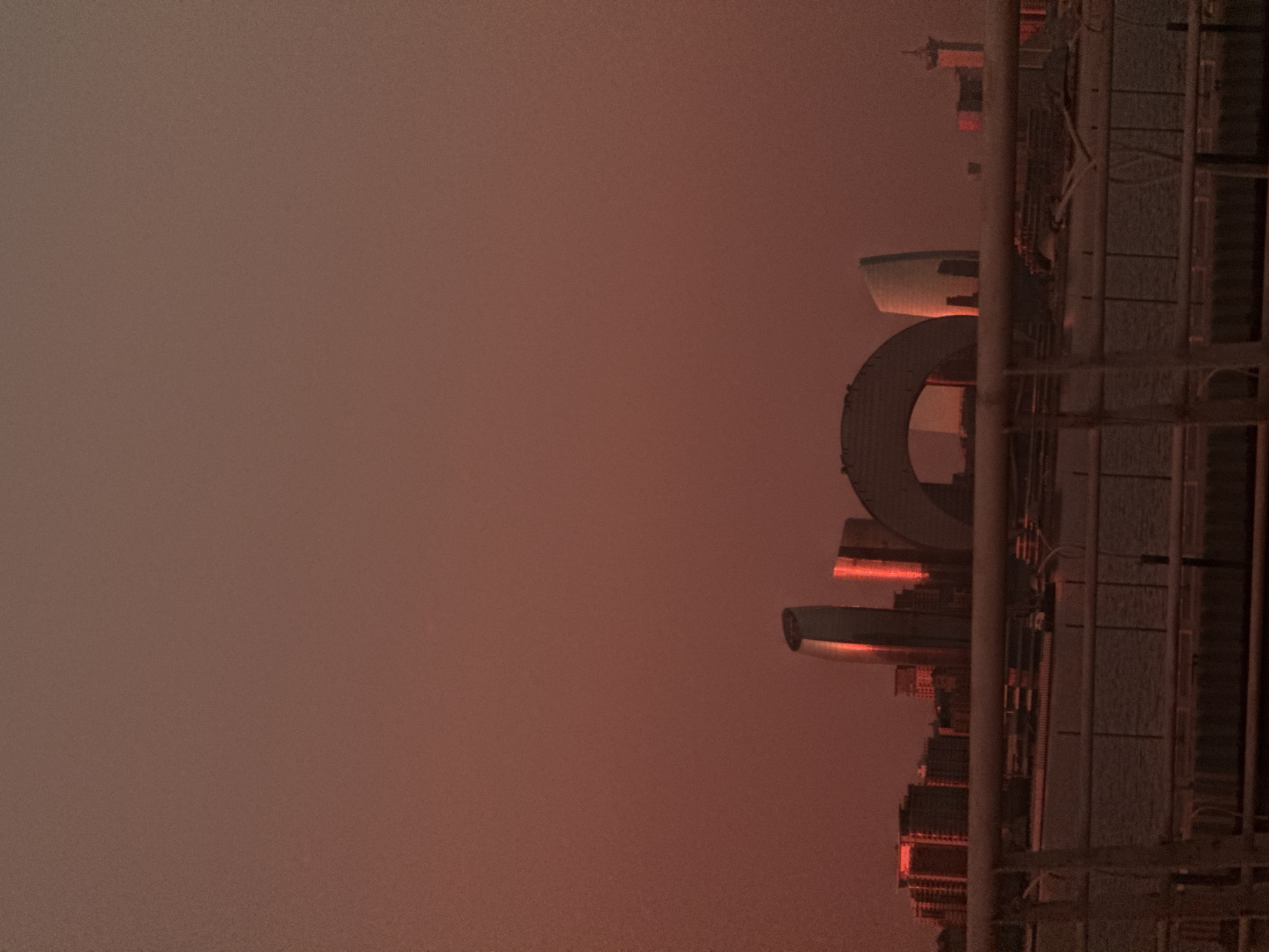The image depicts the Baku Flame Towers and the adjacent modern architecture in Sabail Raion, Azerbaijan, during a sunset or sunrise. The sky is a dominant, uniform reddish-orange hue, suggesting atmospheric conditions or the specific lighting of the sun at a low angle. In the middle ground, the distinctive flame-shaped Flame Towers are visible, their glass facades reflecting the intense red light of the sky, making them appear to glow. To the left and right of the Flame Towers, other contemporary buildings form a city skyline. A large, arched structure, possibly part of an entertainment complex or landmark, is also present in the center. In the foreground, a body of water, likely the Caspian Sea, stretches across the lower portion of the image. A metal railing or fence runs horizontally in the lower part of the frame, indicating a viewpoint from a waterfront promenade or structure. The overall scene is one of urban development set against a dramatic natural sky. No people are visible in the image.