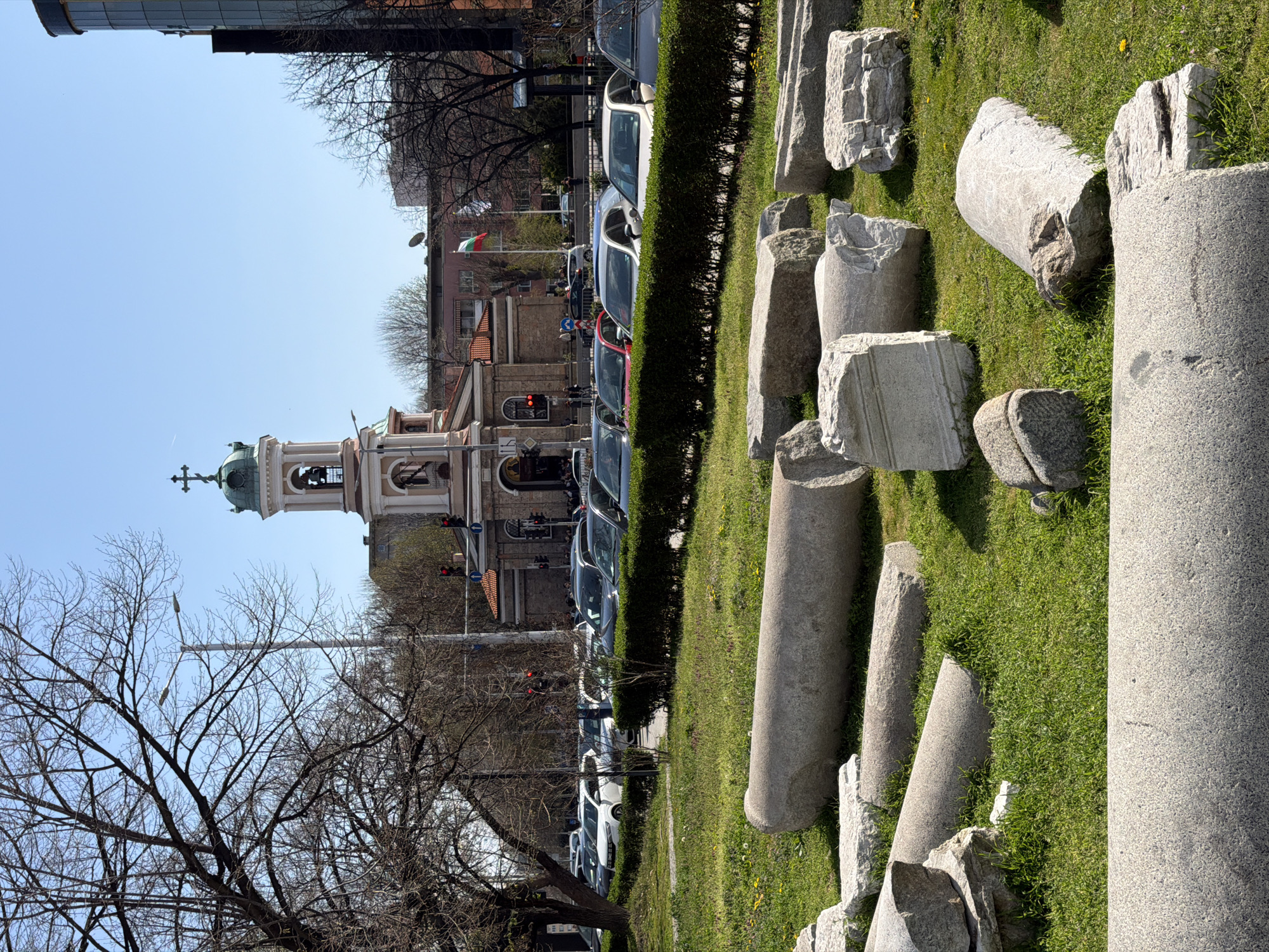 The image shows a view of the Church of St. George in Plovdiv, Bulgaria, with ancient Roman ruins in the foreground. The church, with its distinctive bell tower and green dome topped with a cross, stands on the left of the frame. A Bulgarian flag is visible on a nearby building. Between the church and the ruins, cars are parked alongside a hedge, indicating a street. The ruins consist of broken columns and stone fragments scattered on a grassy area. A bare tree occupies the lower left corner of the image. The sky is blue and clear.