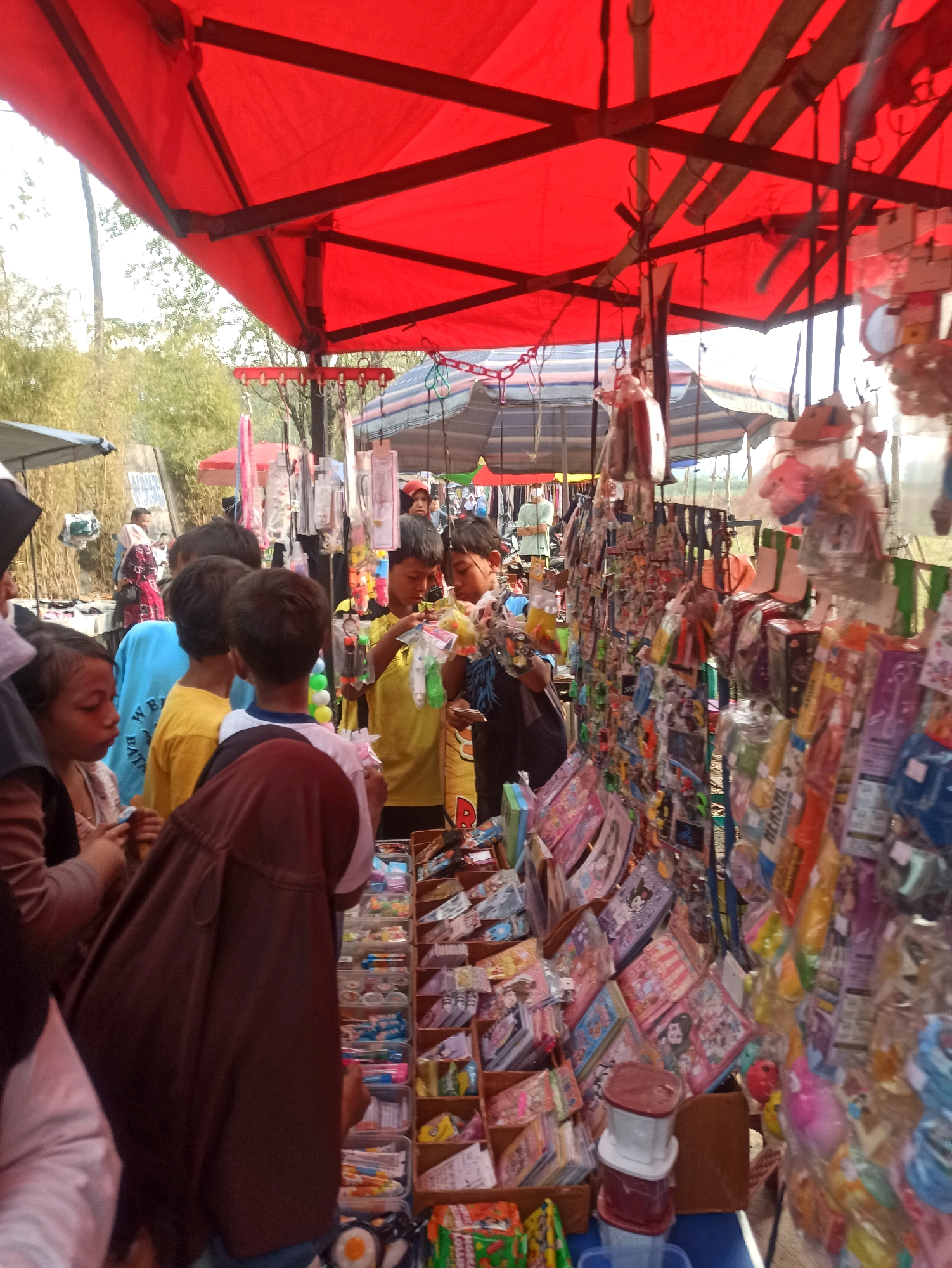 This image depicts a lively outdoor market stall, possibly located in Telagamurni, Indonesia. A vibrant red canopy provides shade over the scene, where various goods are displayed for sale. Several children are gathered, some looking at the merchandise, others holding items. The stall is filled with a variety of products, including packaged candies, toys, and small trinkets, many of which are brightly colored and individually wrapped. The background suggests a sunny day with trees and other market stalls visible. The overall impression is one of bustling commerce and youthful excitement.