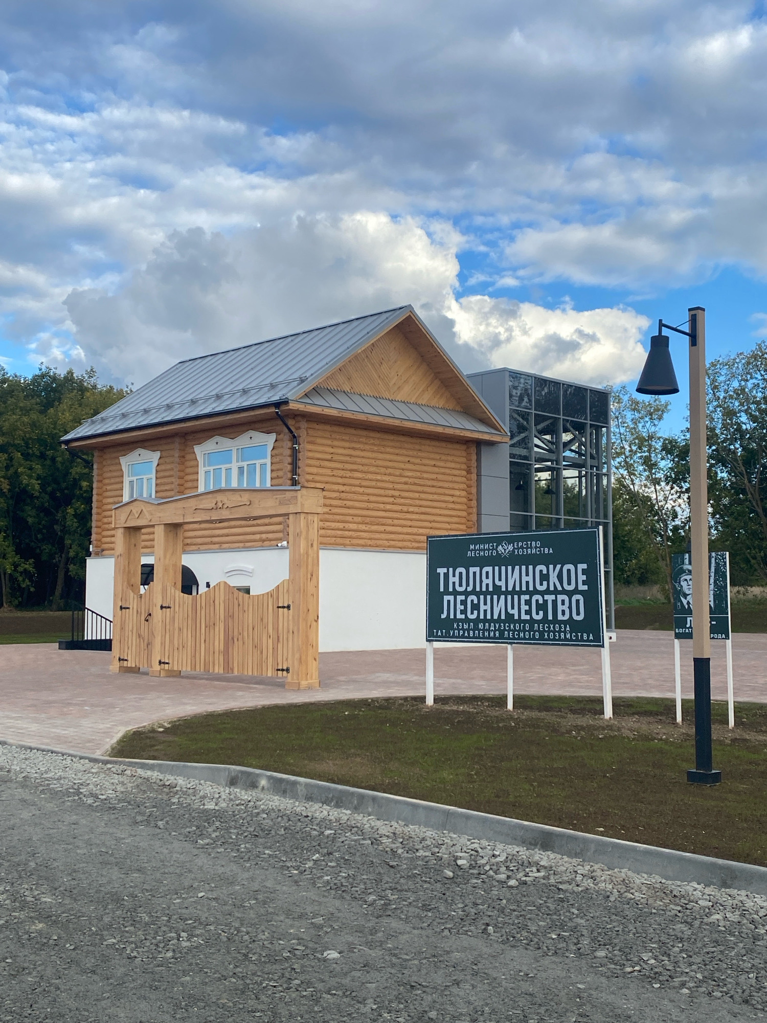 The image shows a two-story wooden building with a metal roof, located in Gorokhovoe Pole, Russia. The building has white-trimmed windows and a wooden gate in front of an entrance. Adjacent to the wooden building is a modern glass structure. In the foreground, there are two green signs with white lettering, one larger than the other, positioned on white poles. A black lamppost stands to the right of the signs. The ground in front of the building is paved with reddish-brown bricks, and beyond that, there is a patch of grass and trees. The sky is cloudy with patches of blue.