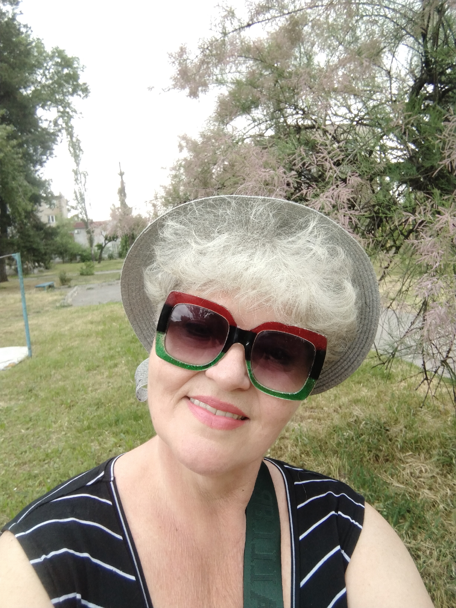 A cheerful woman poses for a close-up selfie outdoors in Horishni Plavni, Ukraine. She is shown from the chest up, looking directly at the camera with a bright smile. Her short, curly, light-colored hair, possibly white or very light blonde, is partially visible beneath a light grey, woven hat. She wears large, fashionable sunglasses with distinctive frames that are red on the upper half and sparkling green on the lower half, featuring dark gradient lenses. Her lips are painted with a light pink lipstick.
The woman is dressed in a sleeveless black top with thin white horizontal stripes. A dark green strap, likely from a bag or another garment, can be seen over her right shoulder.
The background depicts a green, grassy area, suggesting a park or public garden. To her left, there are leafy green trees and a distant multi-story building. A light blue metal structure, possibly playground equipment, is also visible in the mid-ground. To her right, a tree with delicate, feathery, light pink or purplish blossoms or foliage (resembling a Tamarix) fills the upper right portion of the frame. The sky above is bright and appears overcast or hazy. The image captures a pleasant daytime moment. There is no discernible text visible.