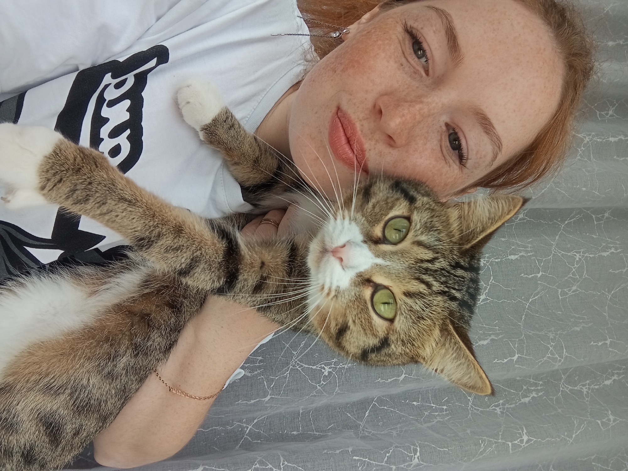 A woman with freckles and red hair lies on a grey marbled surface, wearing a white t-shirt with a black graphic on it. She is holding a brown tabby cat with white paws and green eyes. The location is not specified in the image and there is no way to infer it from the provided data.