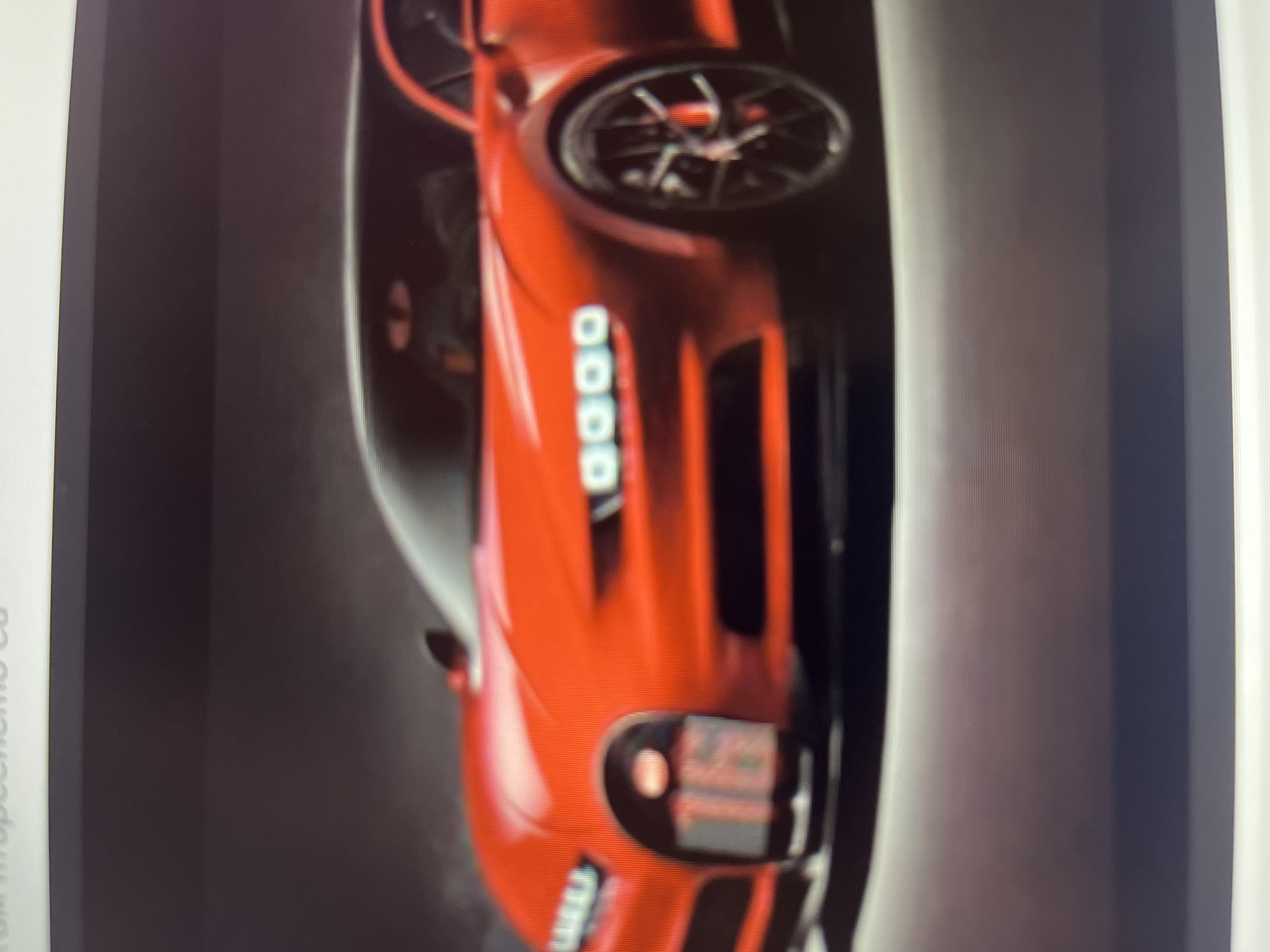 The image displays a red sports car, oriented vertically, with its front section visible. The car appears to be in a dark, studio-like setting, possibly a digital render or a professional photograph. Notable features include multi-spoke wheels, distinctive LED strip headlights, and a prominent front grille. The car's surface is glossy, reflecting light. The overall scene suggests a promotional or display context for the vehicle, which is stationary. The image itself appears to be a photograph taken of a screen, exhibiting some blur and faint scan lines. On the far left edge of the original image, a partial and blurry text string is visible, starting with "um troopesome ca" or similar, indicating a possible watermark or website address, but it is largely illegible.