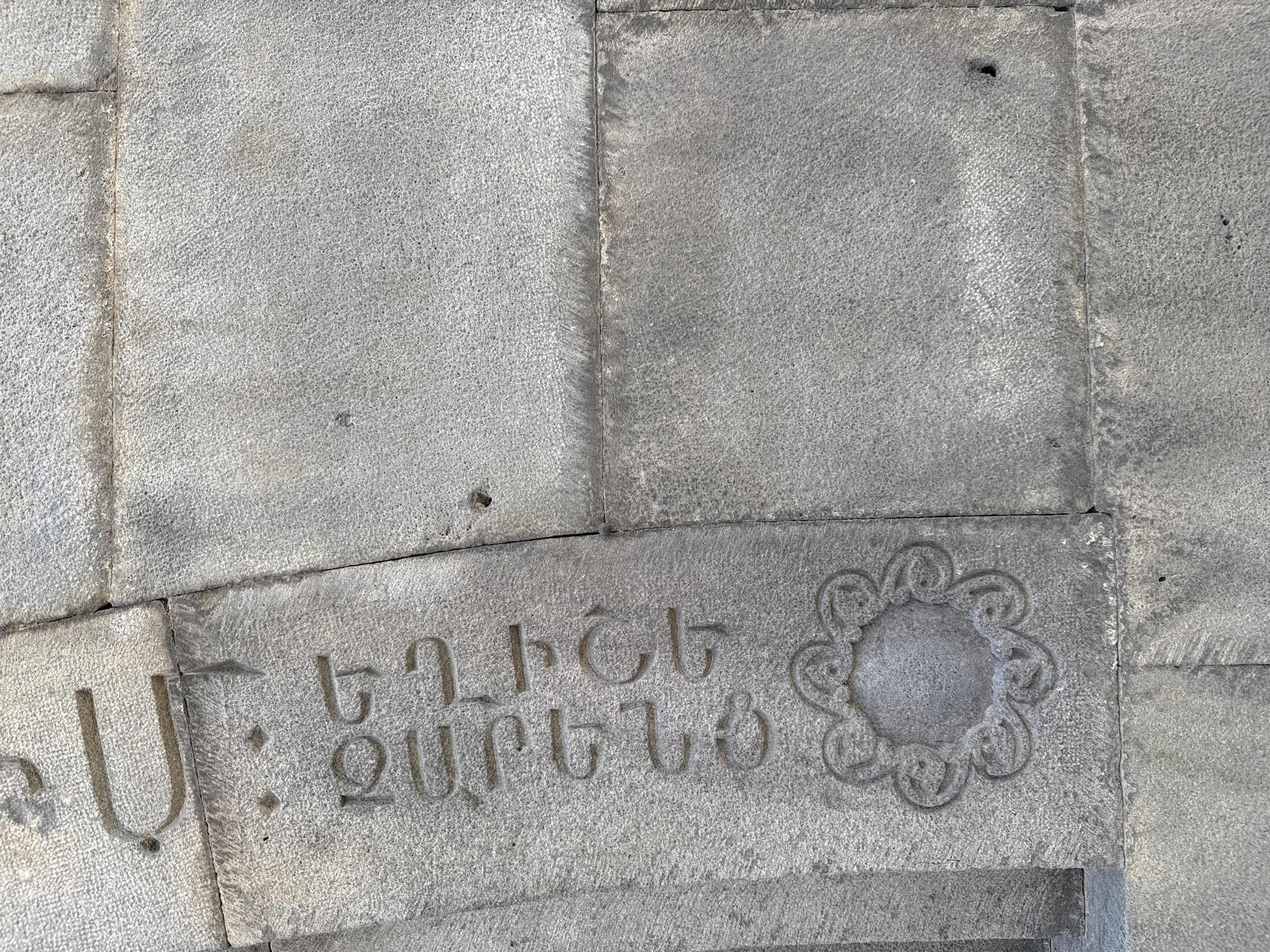 The image shows a close-up view of weathered stone carvings on a building facade. The stone is a light grey, textured granite, with visible mortar lines separating the blocks. The carvings appear to be in an ancient script, possibly Armenian, with several vertical lines of text and a decorative floral motif at the bottom left. The script is incised into the stone, with some letters showing wear and erosion. The floral design is circular, with intricate swirling patterns. The overall impression is one of age and historical significance. The location is identified as Voghjaberd, Armenia, suggesting this is part of a historical structure or monument in that region. There are no people or animals visible in the image. The lighting is natural, casting subtle shadows that highlight the texture of the stone and the depth of the carvings.