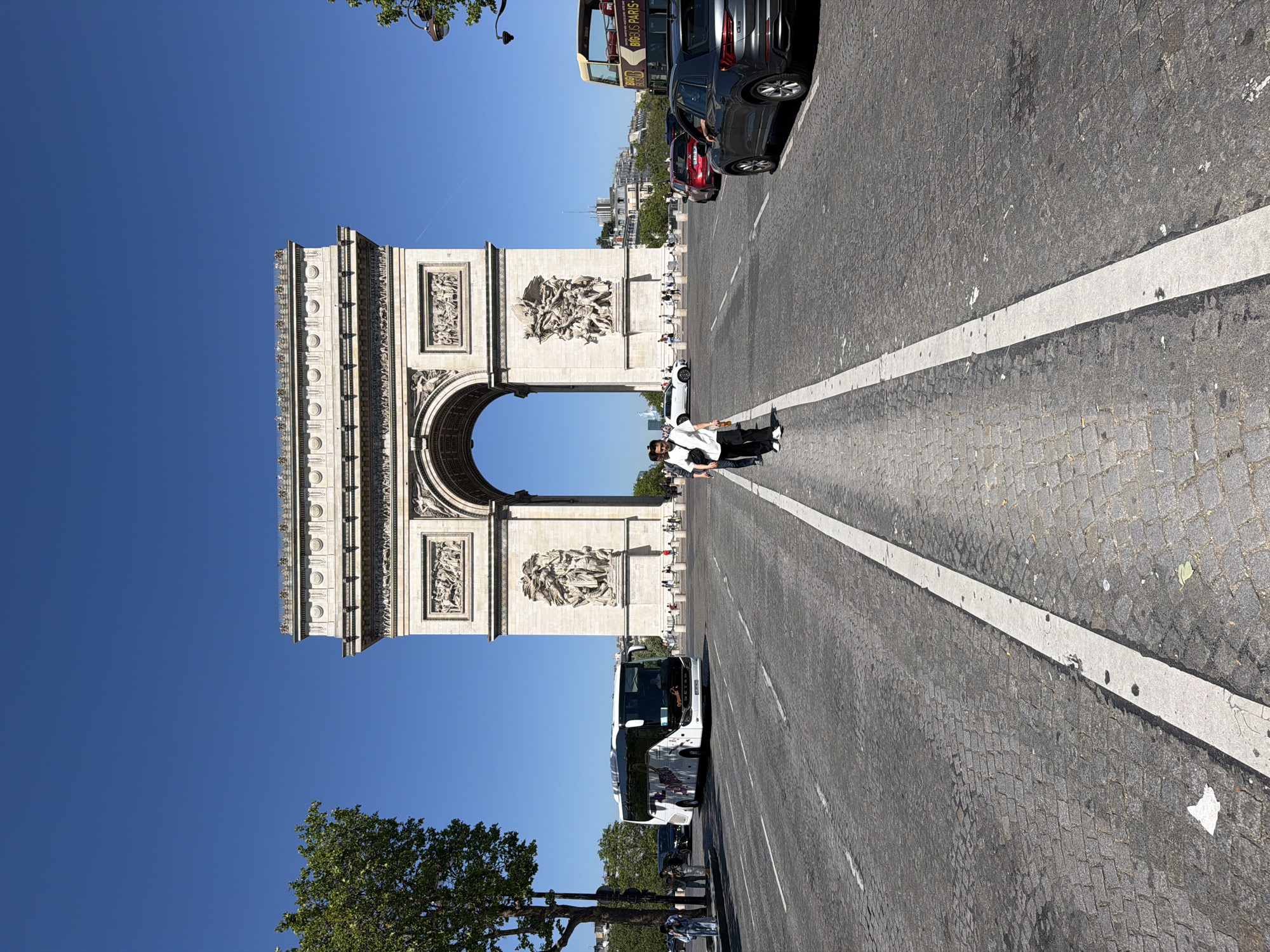 The image shows the Arc de Triomphe in Paris, France. The arch is in the background. In the foreground, a cobblestone street stretches out towards the arch. There is a solid white line marking the center of the road. Several cars and buses are visible on the right side of the image. Two people are standing in the street, near the center line, appearing to pose for a photo. A tree is visible in the lower left corner. The sky is clear and blue.