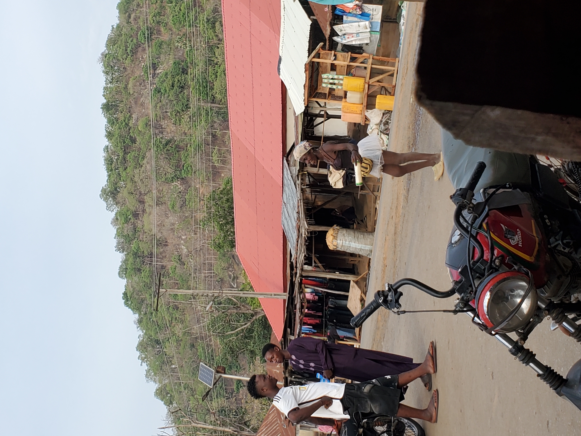 The image captures a vibrant street scene in Mararaba, Nigeria, situated at the base of a lush, tree-covered hillside under a bright, clear sky. Power lines are visible running along the base of the hill and across the landscape.
In the foreground and midground, several individuals are engaged in daily activities. On the left, two men are conversing beside a motorcycle and a pole supporting a small solar panel. One man wears a white t-shirt and dark shorts, while the other is dressed in a long, dark purple garment. To the right, another motorcycle is partially visible, featuring a round headlight and the text "HONDA" on its side.
Further down the unpaved road, towards the right, a woman stands near a row of market stalls constructed with wooden frames and corrugated metal roofs. She wears a light top, a skirt, and a head covering, appearing to be attending to merchandise. The stalls display various bags and bright yellow jerrycans, suggesting the sale of goods or water. In the background, behind the market stalls, more buildings with prominent red corrugated roofs are visible, blending into the natural environment. The scene depicts typical daily commerce and interactions in a sunny, semi-urban or rural setting.