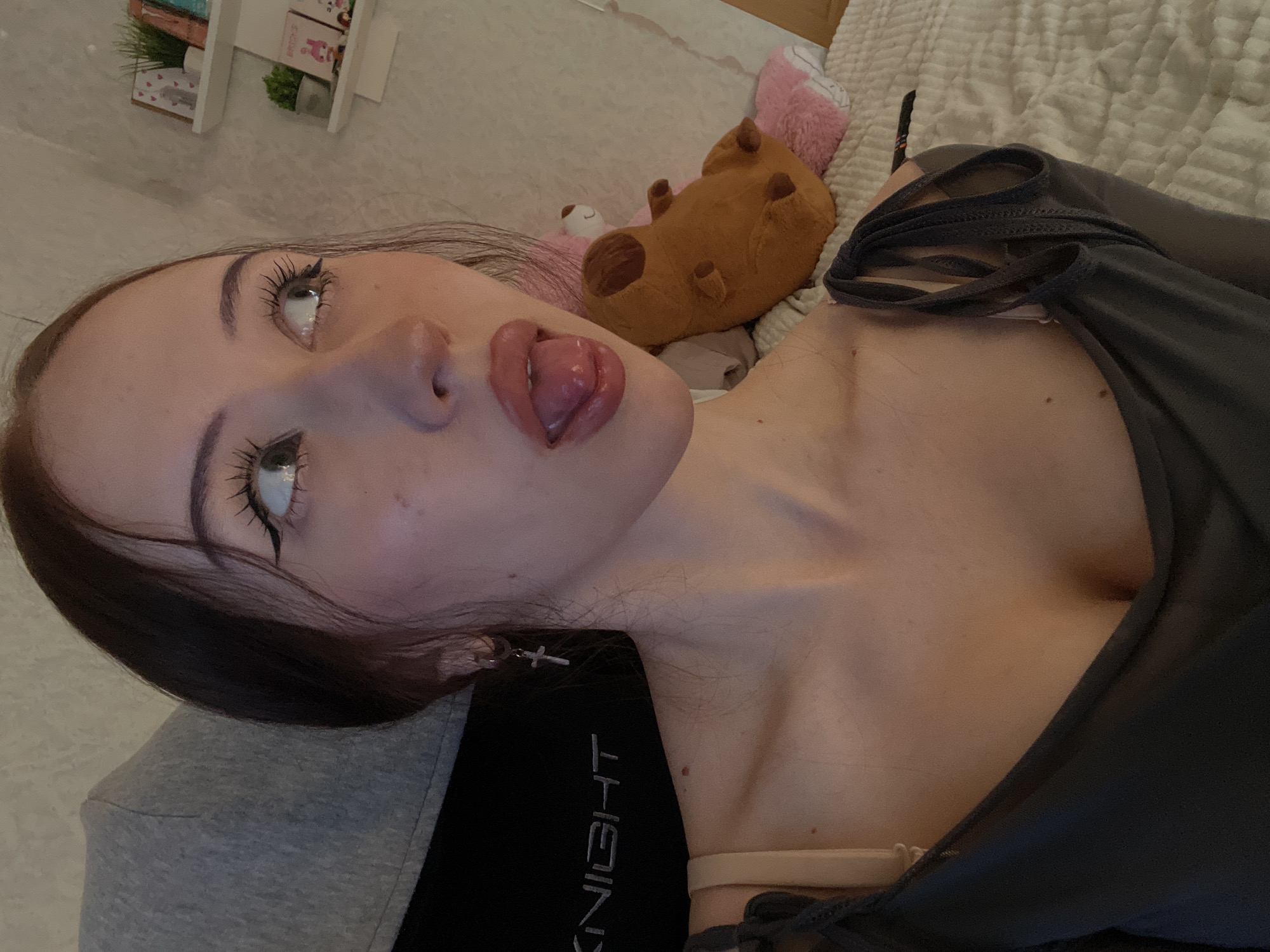 A young adult female is depicted in an indoor setting, visible from the upper chest upwards. She is positioned with her head slightly tilted and turned to her right, gazing upwards and to her right, with her tongue partially extended from her mouth. She has dark hair, styled with some strands framing her face, and prominent winged eyeliner and mascara are visible on her eyes. Her lips appear full and glossy. She wears a dark grey top with visible strap detailing on her right shoulder, and a light beige bra strap is visible on her left shoulder. A silver hoop earring with a small silver cross pendant hangs from her left earlobe. A small, dark mole is present on her upper chest, near her clavicle.
Behind her head to the left, a grey fabric material is visible, possibly belonging to a hooded garment or a chair. Below this, a black surface with the word "KNIGHT" in reflective capital letters is partially discernable, suggesting part of a gaming chair or similar furniture. To her right, a light-colored wall with a subtle textured finish is visible. Two white floating shelves are mounted on this wall. The upper shelf holds a small potted green plant, two framed decorative items (one featuring a red heart), and what appear to be books or small boxes. The lower shelf displays another small potted green plant, items labeled "BRIGHTS," a small pink character figurine, and other decorative objects. In the lower right of the frame, light-colored, textured bedding or a blanket is visible, on which rest two stuffed animals: a brown, dog-like or bear-like plush, and a larger pink plush partially obscured behind it. A thin, dark, elongated object, possibly a remote control or writing utensil, lies on the bedding near her right shoulder. The image contains no visual information to indicate its precise geographical location, including the city of Votkinsk, Russia.