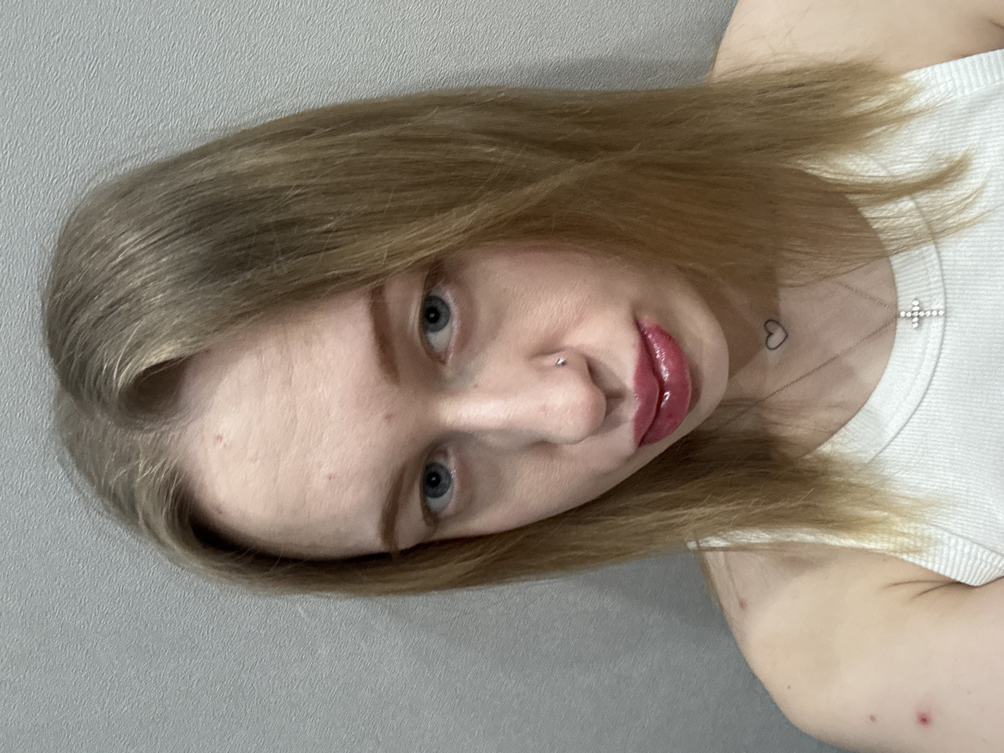 A female individual is depicted in a close-up portrait, located in Rostov-on-Don, Russia. The subject, positioned centrally, has light brown hair, blue eyes, and is looking directly at the camera. She has a small silver stud piercing on her left nostril. Her lips are coated with a glossy, dark pink lipstick, showing minor smudging or irregular application on the lower lip. Visible skin blemishes are present on her forehead, right cheek, and left shoulder/upper arm. She is wearing a white ribbed tank top. A small black outline of a heart is tattooed on the right side of her neck, and a delicate necklace with a small, possibly jeweled, cross pendant rests on her chest. The background consists of a plain, light grey wall with a subtle texture.