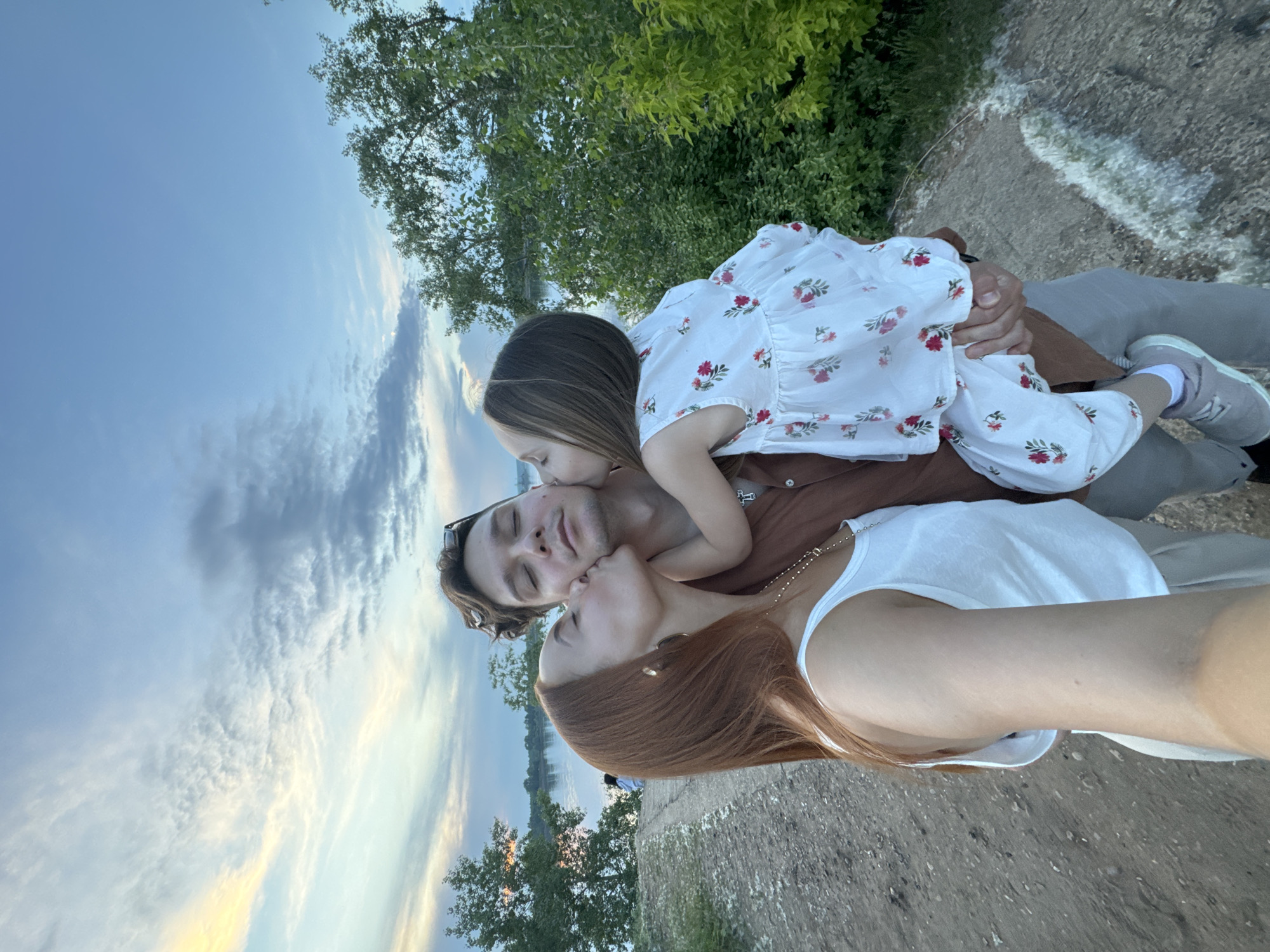 A family of three, a father, mother, and daughter, are captured in a selfie outdoors. The father is in the center, with his eyes closed, receiving kisses from both his wife and daughter. The wife, on the left, has reddish-brown hair and is wearing a white tank top and a necklace. The daughter, in the father's arms, is wearing a white dress with a floral pattern and pink shoes. They are standing on a dirt path with trees and a body of water in the background. The sky is partly cloudy, with hues of blue and orange indicating it may be sunset or sunrise. The location is the embankment of the Kama River in Chaikovsky, Russia.