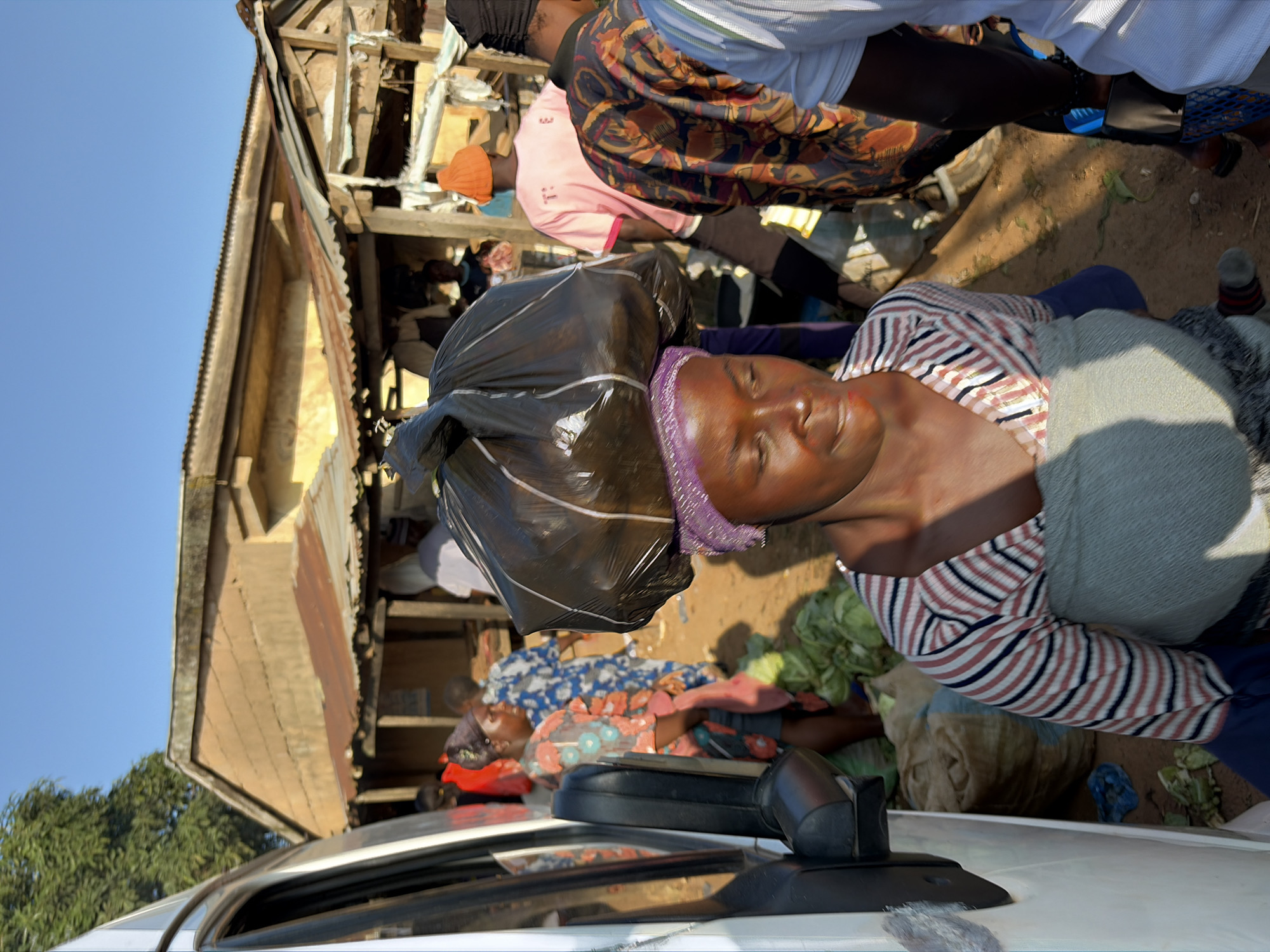 In Gura Top, Nigeria, a woman balances a large black plastic bag of goods on her head while walking through an outdoor market. She wears a striped long-sleeved shirt and a light grey wrap around her chest. Several other people are visible in the background, some carrying goods and others tending to stalls. The environment appears to be dusty with corrugated metal roofs and wooden structures typical of a market. A white van is partially visible on the left side of the image. The sky is clear and blue, indicating daylight.