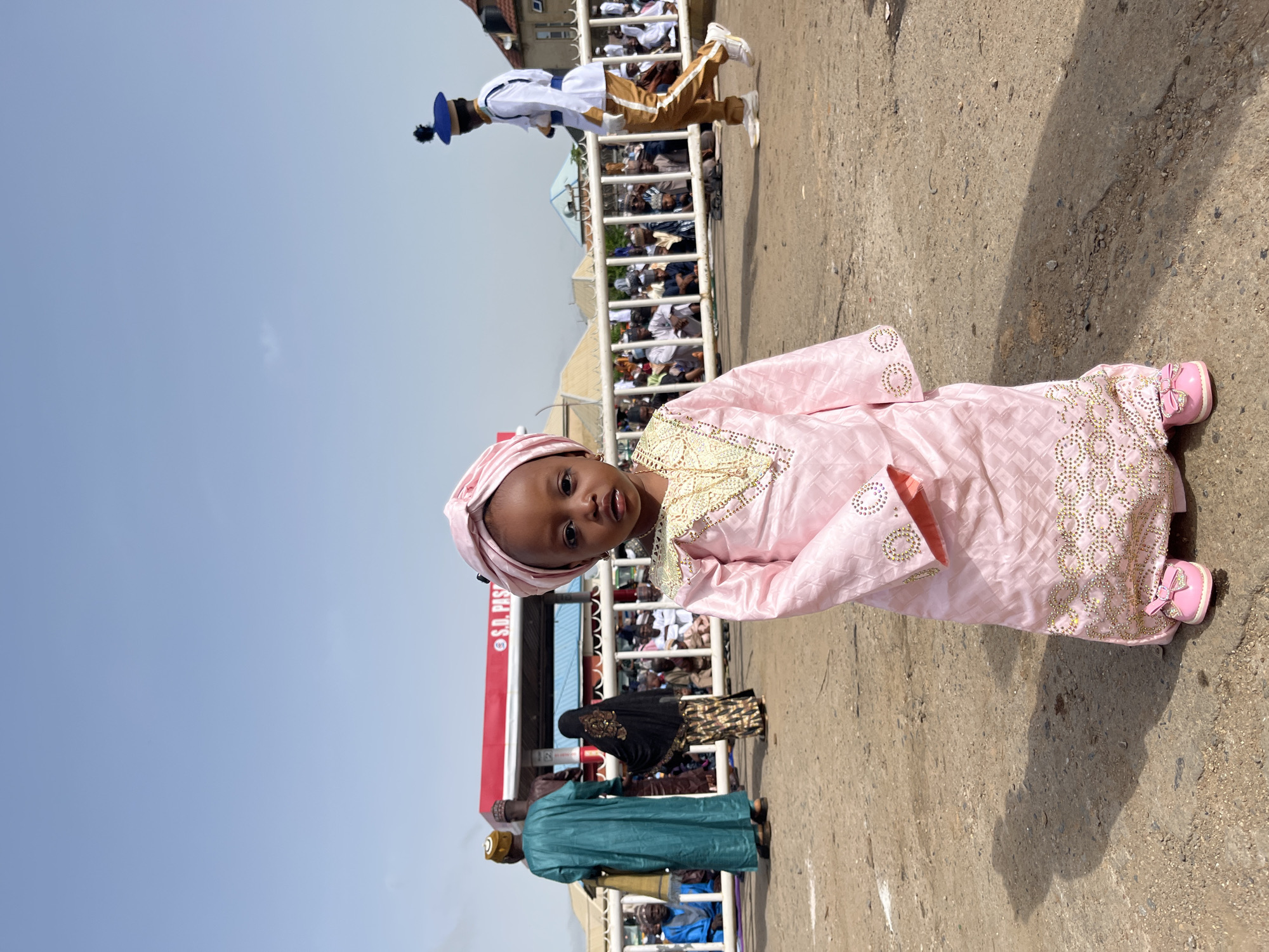 A young child wearing a pink traditional garment and head covering is standing on a concrete surface in what appears to be a public gathering. Behind the child is a white metal barricade separating the crowd from the street. A person in traditional dress is marching or dancing behind the barricade. The location is identified as Jos, Nigeria based on signage in the background that says "S.D. Pass". Other individuals, including some in traditional attire, are visible behind the barricade. The sky is clear and blue.