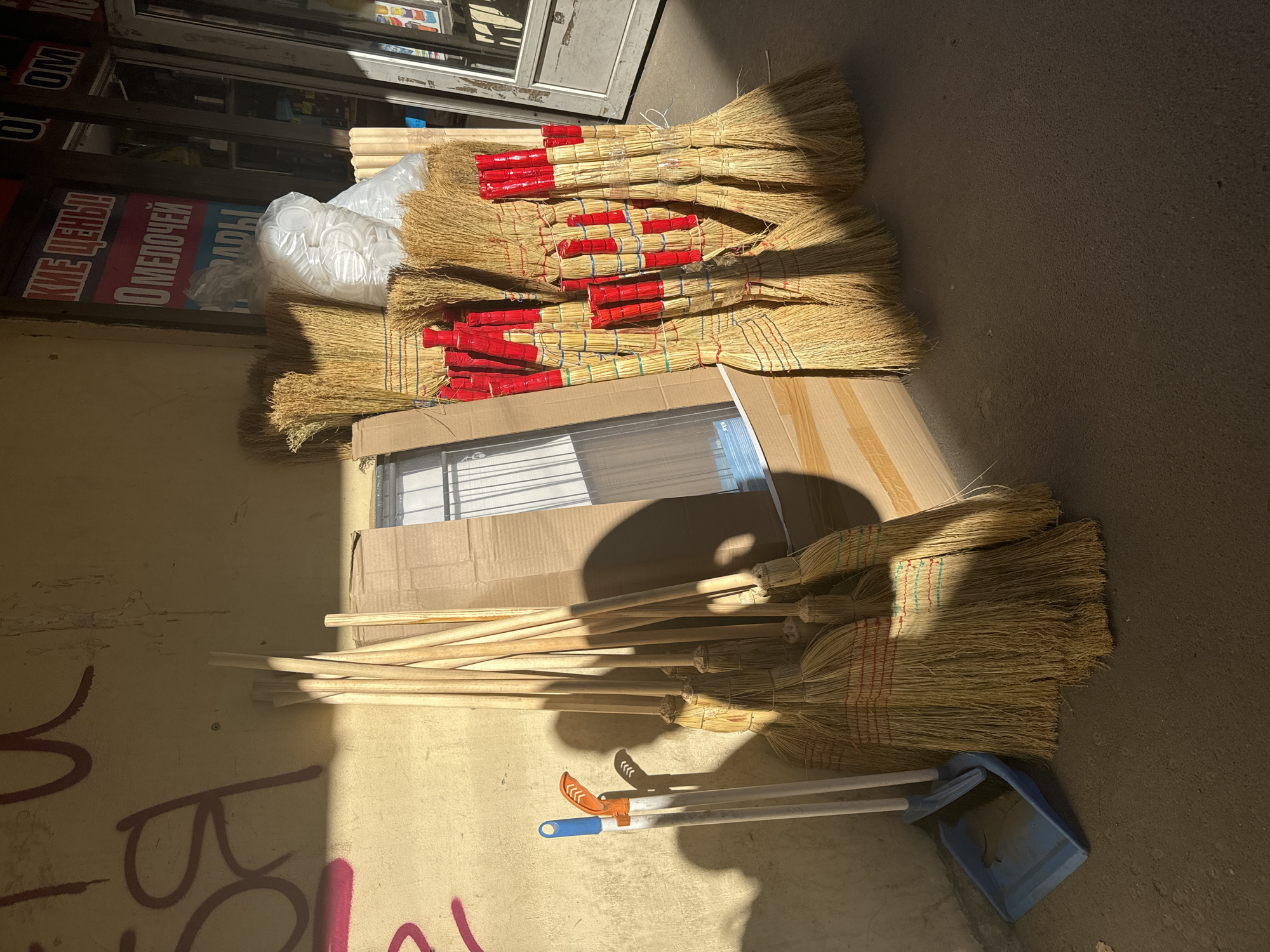 This is a medium-angle, exterior shot of cleaning supplies stacked against a beige concrete wall with graffiti. The scene is outdoors on a sunny day, casting shadows. In the foreground, a blue plastic dustpan and two broom handles (one white and orange, one white) are leaning against the wall. Behind them, a large bundle of straw brooms, their handles made of light-colored wood, are leaning against a cardboard box. The brooms have red and blue twine bindings near their heads. A clear plastic bag filled with white, circular objects is placed behind the brooms. The wall has large, dark red graffiti markings. To the right, a storefront with a glass door and windows is visible, with signage displaying text in Cyrillic. The ground is asphalt. The scene appears to be a street-level storage or display area, likely in a commercial district of Saint Petersburg, Russia.