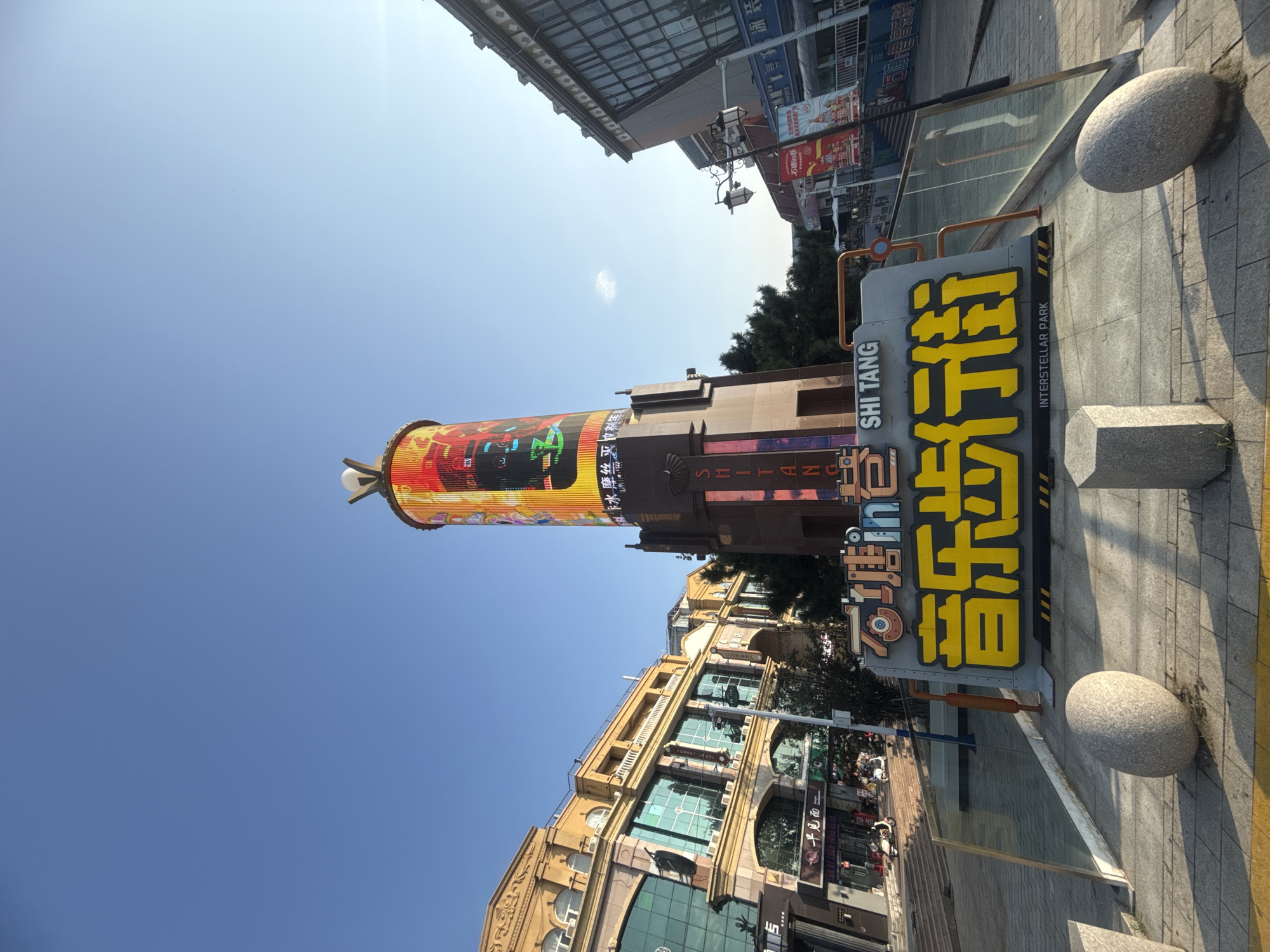 The image shows a wide-angle, eye-level view of a street scene in the city of Qinhuangdao, China, specifically in an area identified by signage as "SHI TANG INTERSTELLAR PARK" and featuring a prominent sign with Chinese characters for "Music Street" in large yellow lettering. The focal point of the image is a tall, cylindrical tower with a digital display showing colorful graphics and text. To the left of the tower is a building with a tan facade, arched walkways on the ground floor, and large green-tinted windows. To the right of the tower, another building with a darker facade and a mansard roof is visible. The foreground features a paved walkway with two large, smooth, oval-shaped stones and a rectangular stone block. A glass barrier runs along the edge of the walkway. The sky is clear and blue, with the sun casting a warm light on the scene, suggesting it is either morning or late afternoon. No individuals are clearly distinguishable in the image, though there are some faint figures on the street level in the distance, and some parked scooters. The overall impression is of an urban commercial or entertainment district.