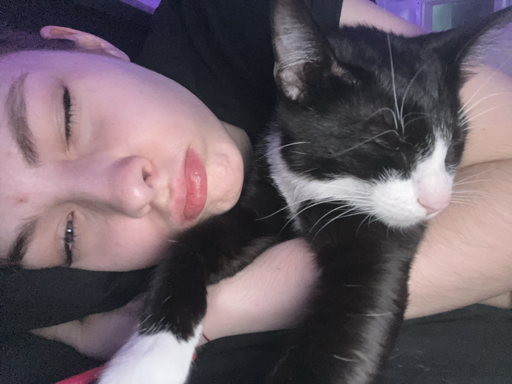 A close-up photograph depicts a person and a black and white cat in close proximity. The person is fair-skinned with their eyes partially open. The cat is positioned closely to the person, appearing to be asleep or resting comfortably. The background is dark and indistinct. The location of this scene is not apparent from the image.