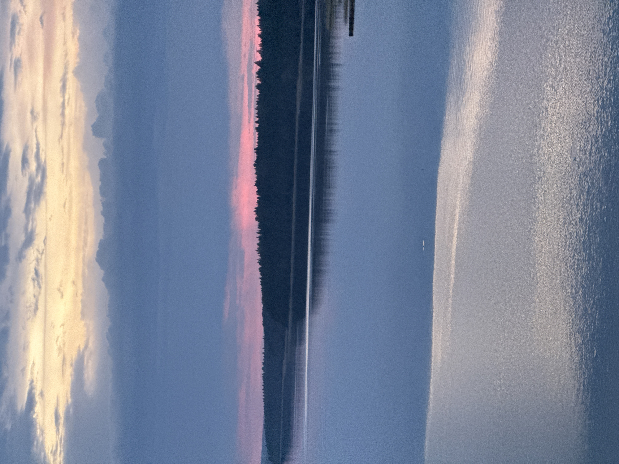 The image depicts a body of water, possibly a lake or wide river, with reflections of the sky and shoreline. On the left, there is a sunset with yellow and white clouds blending into a blue sky. In the middle, a dark treeline separates the water from the sky, with a pinkish glow indicating the setting sun. A thin, vertical structure is visible above the treeline on the right side. The water's surface is generally calm. The location is not specified but mentioned the city of Votkinsk, Russia, which is not visibly indicated.