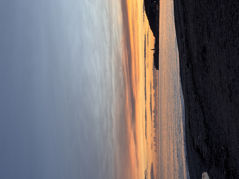 This media file presents a serene coastal landscape captured during either sunrise or sunset, as indicated by the vibrant colors of the sky. The image is oriented vertically but depicts a horizontal scene.
In the upper portion of the frame, the sky dominates, displaying a spectacular gradient. On the left, it begins with a soft, cool grey-blue, transitioning towards the right into warmer tones of pale yellow, orange, and hints of pink. Wispy, elongated clouds stretch across the sky, catching and diffusing the light, creating a painterly effect.
Below the horizon line, which is approximately in the middle of the vertical frame, lies a calm body of water, likely the sea. This water beautifully reflects the warm colors of the sky, shimmering with golden and orange hues.
Along the horizon, a dark silhouette of a coastline or landmass is visible. This land features the outlines of structures, including what appears to be a prominent church spire, strongly suggesting the Old Town of Budva, Montenegro, clinging to the edge of the Adriatic Sea.
The lower portion of the image shows a dark, textured area, characteristic of a rocky beach or dark sand/pebbles, providing a stark contrast to the bright sky and reflective water. The texture is coarse and uneven, implying a natural, undeveloped shoreline.
The scene conveys a sense of tranquility and natural beauty, with no human activity or individuals present. The time of day is clearly either dawn or dusk, creating a peaceful and picturesque atmosphere. There is no visible text within the image.