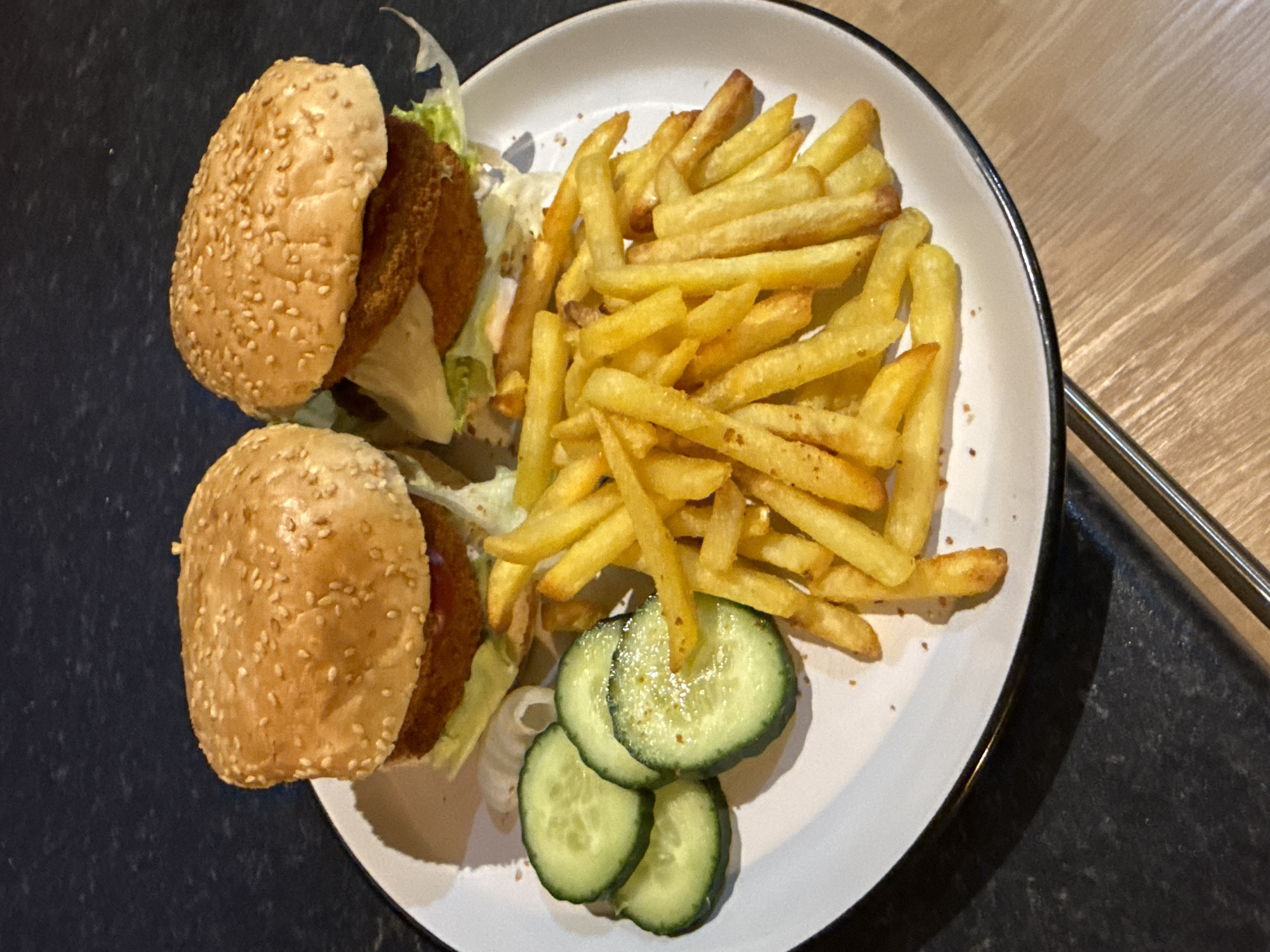 The image shows a plate of food containing two small burgers, fries, and cucumber slices. The burgers have sesame seed buns, lettuce, and a patty. The fries are yellow and appear to be lightly seasoned. There are three cucumber slices. The plate is white with a black rim and is placed on a surface that is part black and part wood. Silverware is seen to the right of the plate. The location is not discernable from the image. There is no indication the location is in the city of Great Yarmouth, United Kingdom.