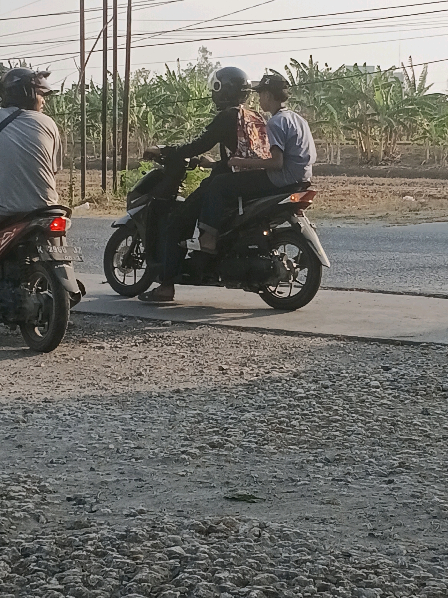 Two individuals are riding on a black motorcycle. The person in front, wearing a helmet and a dark top, is operating the motorcycle. The person behind, also wearing a helmet, is wearing a blue t-shirt and has their arms around the driver. A second motorcycle is visible on the left, ridden by a person wearing a grey t-shirt and a helmet, who is facing away from the camera. The motorcycles are on a paved road, with a gravel shoulder on the right. In the background, there are utility poles and a dense plantation of banana trees. The scene appears to be in a rural or semi-rural area of Purwodadi, Indonesia, based on the vegetation and architecture. The lighting suggests it is daytime, possibly late afternoon.