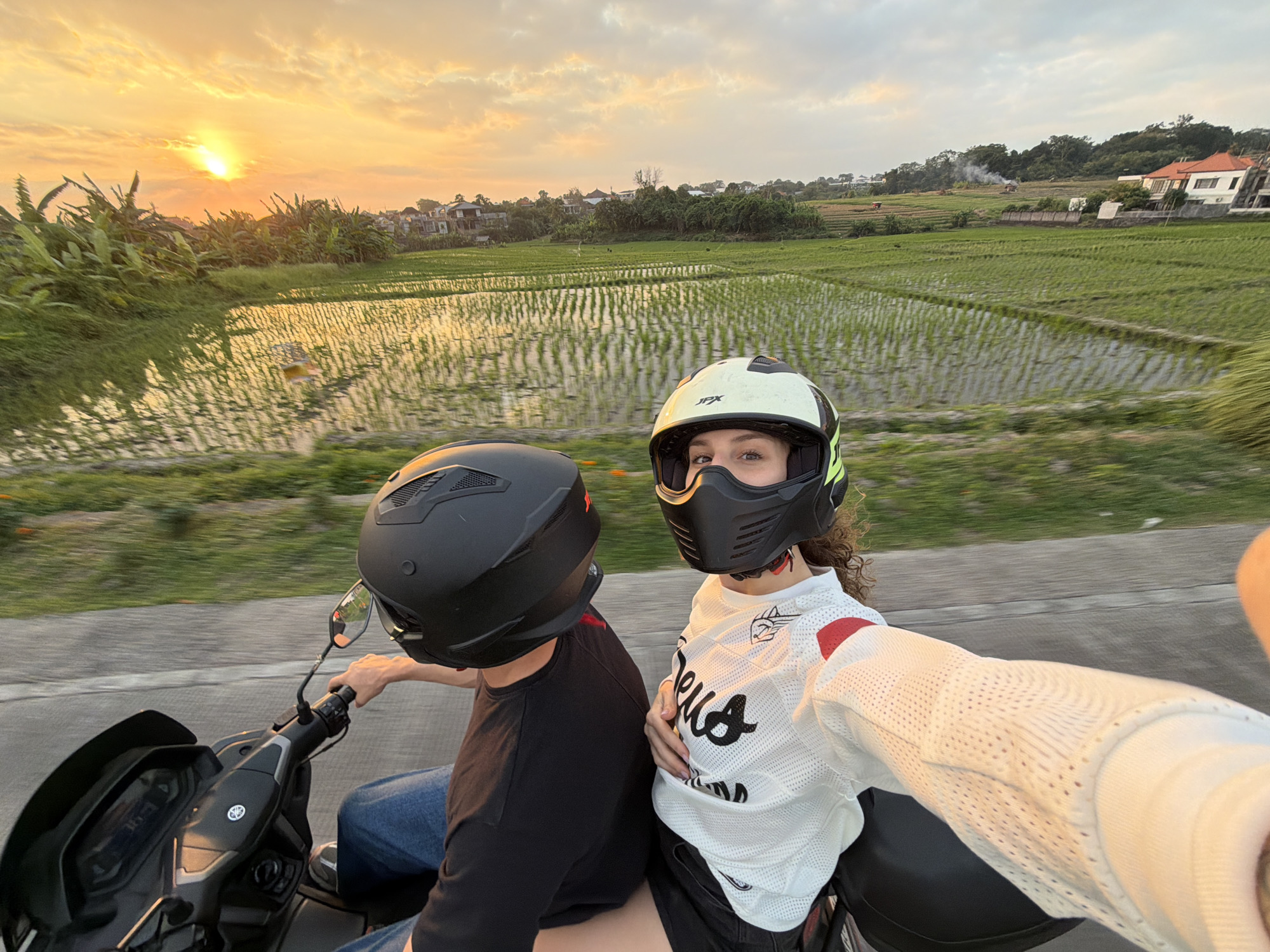 Two individuals are riding a motorcycle through a rural area in North Kuta, Indonesia, during sunset. The person in the foreground, wearing a white long-sleeved shirt with red accents and a black and white helmet, is holding the camera and appears to be taking a selfie. They have curly brown hair and are looking at the camera. The person behind them, wearing a black t-shirt and jeans with a black helmet, is driving the motorcycle. The motorcycle is black and has a Yamaha logo on the front.
The landscape features lush green rice paddies reflecting the golden light of the setting sun. Banana trees and other vegetation line the side of the road, with houses visible in the background. The sky is filled with warm orange and yellow clouds, indicating the sun is low on the horizon. The road appears to be paved. The motion blur suggests the motorcycle is in motion.