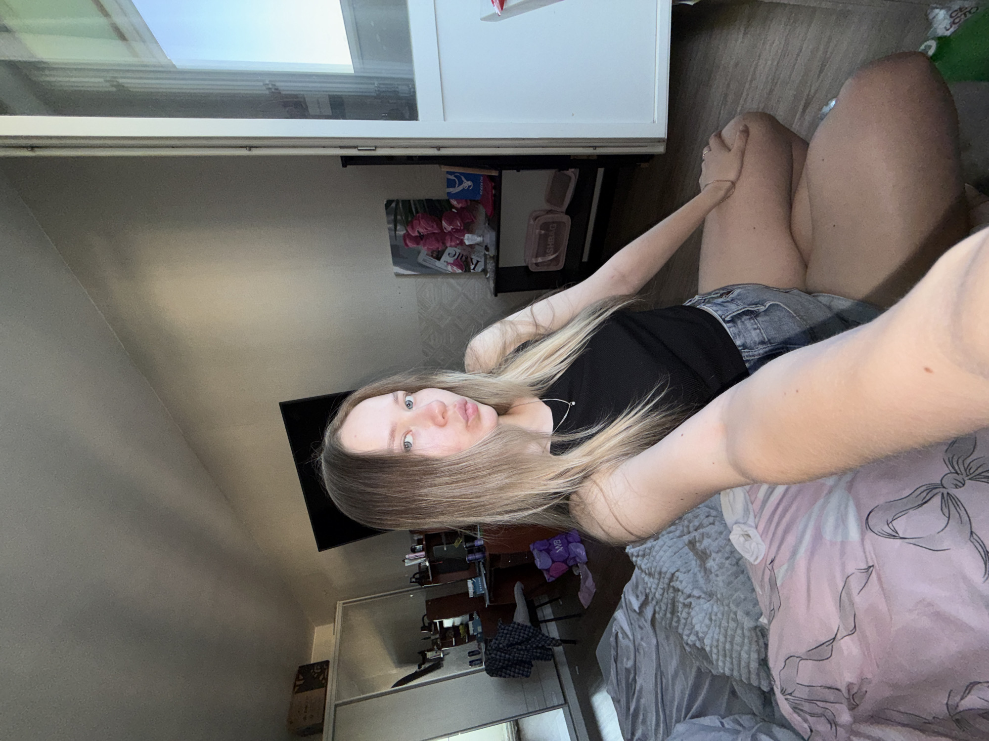 A woman is in a room. She is on a bed with a pink comforter and a gray blanket. She has blonde hair and blue eyes. She is wearing a black tank top and denim shorts. In the background, there is a television, a desk with shelves, and a white door. The location is likely Chaikovsky, Russia, given the request.
