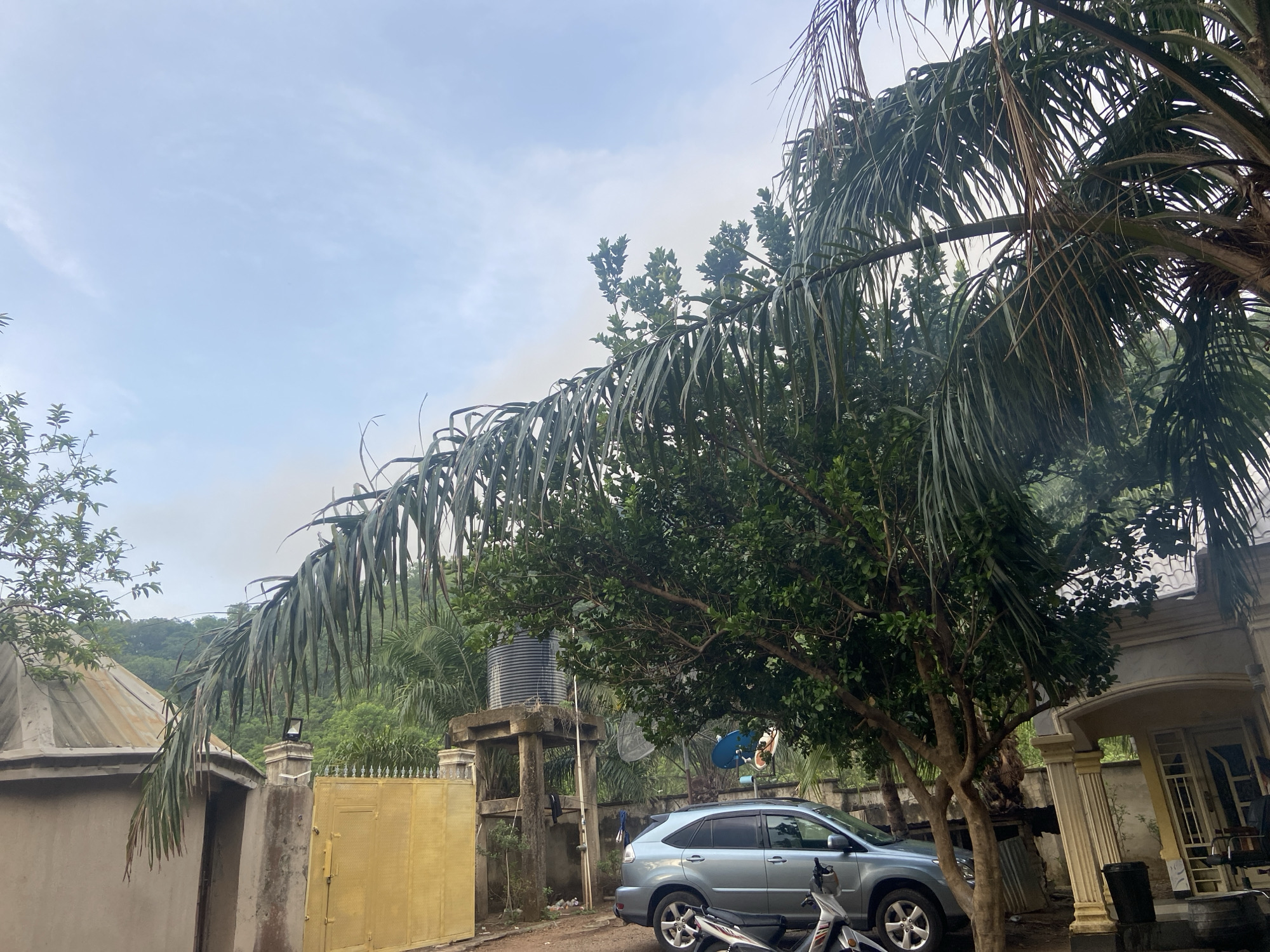 The image presents a street scene in Mararaba, Nigeria. A blue SUV is parked near a residential building with white pillars and beige walls. A motorcycle is parked in front of the SUV. A yellow gate stands next to a concrete water tank supported by pillars. Lush green vegetation and palm trees surround the buildings. The sky is overcast. No people are clearly visible. A satellite dish is attached to a structure near the water tank.