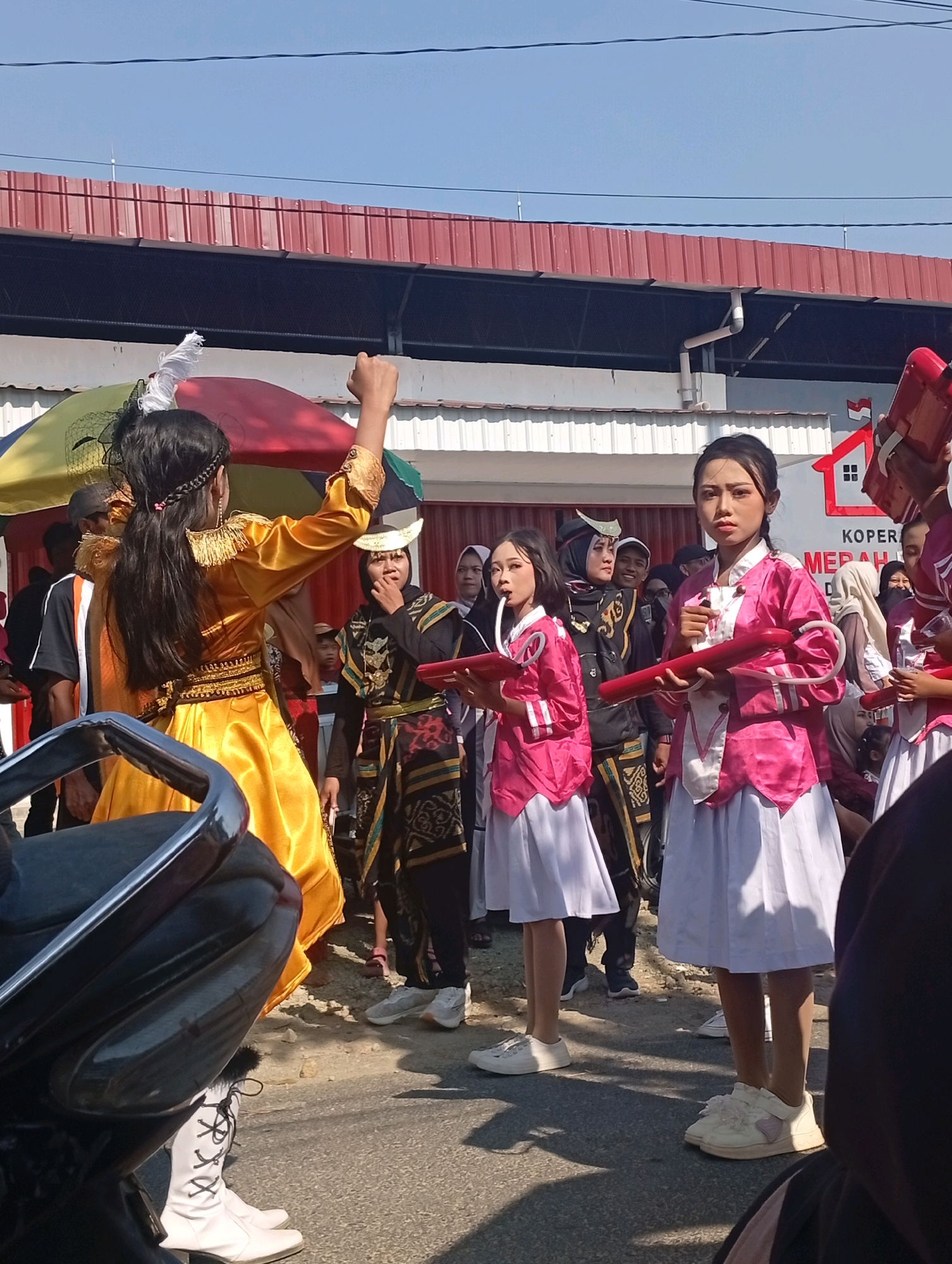 In a street scene in Nglojo, Indonesia, a group of individuals, predominantly young women and some men, are participating in what appears to be a parade or cultural performance. In the foreground, a person in a bright yellow, traditional-looking outfit with intricate gold embellishments is striking a pose with their right arm raised. Their back is mostly towards the camera, but their head is turned slightly to the left.
To their right, two young women are positioned closely together. The one on the left is wearing a pink jacket over a white top and a white skirt, and is holding a red, horn-like instrument to her mouth, as if playing it. The young woman to her right is similarly dressed in a pink jacket and white skirt, and is holding a red, horn-like instrument horizontally in front of her. Their expressions are serious and focused.
Behind these individuals, a diverse crowd is visible. Some are in traditional Indonesian attire, including dark garments with gold patterns and headwear. Others are dressed more casually. A man in a black shirt with an orange stripe is standing to the left of the person in yellow. In the background, buildings with red corrugated roofs are visible, suggesting an urban or semi-urban environment. The sky is clear and blue. The ground is paved with asphalt, and there are some shadows cast by the people and objects present. A portion of a motorcycle is visible in the lower left corner of the frame. The overall atmosphere suggests a lively event taking place outdoors under a bright sun.