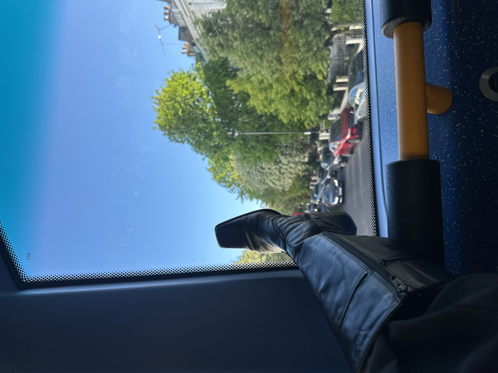 The image captures a first-person perspective from inside a vehicle, likely a bus, looking out through a large window. In the immediate foreground, a person's leg is prominently displayed, with the foot propped up against the bottom frame of the window. The person is wearing a black leather boot with a square toe and a visible zipper running along its side.
The interior of the vehicle shows parts of a blue, speckled upholstered surface on the right, suggesting a seat or interior wall, and a yellow grab rail with black padded sections. The window frame itself has a dark, possibly rubberized, textured strip along its bottom edge, featuring a pattern of small black dots. Small, faint text "V2A" is visible on the lower left of the window glass.
Through the window, a bright, clear blue sky dominates the upper portion of the view, indicating a sunny daytime. Below the sky, the landscape reveals a street scene. Lush green trees, some densely packed, line the street, with multi-story buildings of traditional urban architecture visible beyond them. Numerous cars, including red and white models, are visible on the road, suggesting an urban or suburban setting, possibly experiencing traffic or parked along the street in London, United Kingdom. The overall impression is one of a relaxed journey on a bright day.